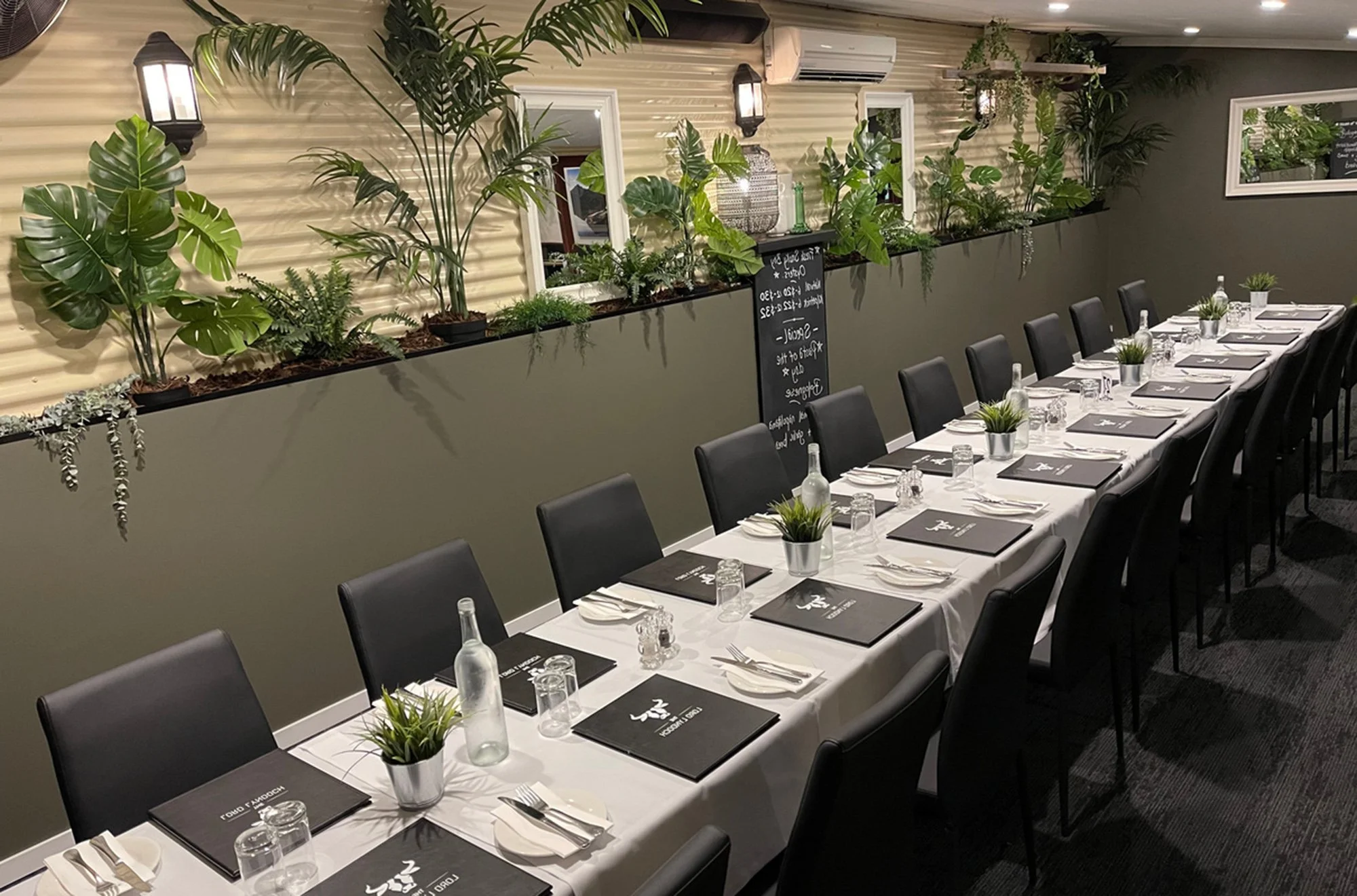 Long dining table set for a meal in a restaurant, with black chairs, white tablecloth, water bottles, glasses, and small potted plants. The background features a wall with various potted plants and a chalkboard menu.