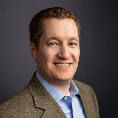 Tyler Huguley smiling in a gray suit and light blue shirt against a dark background.