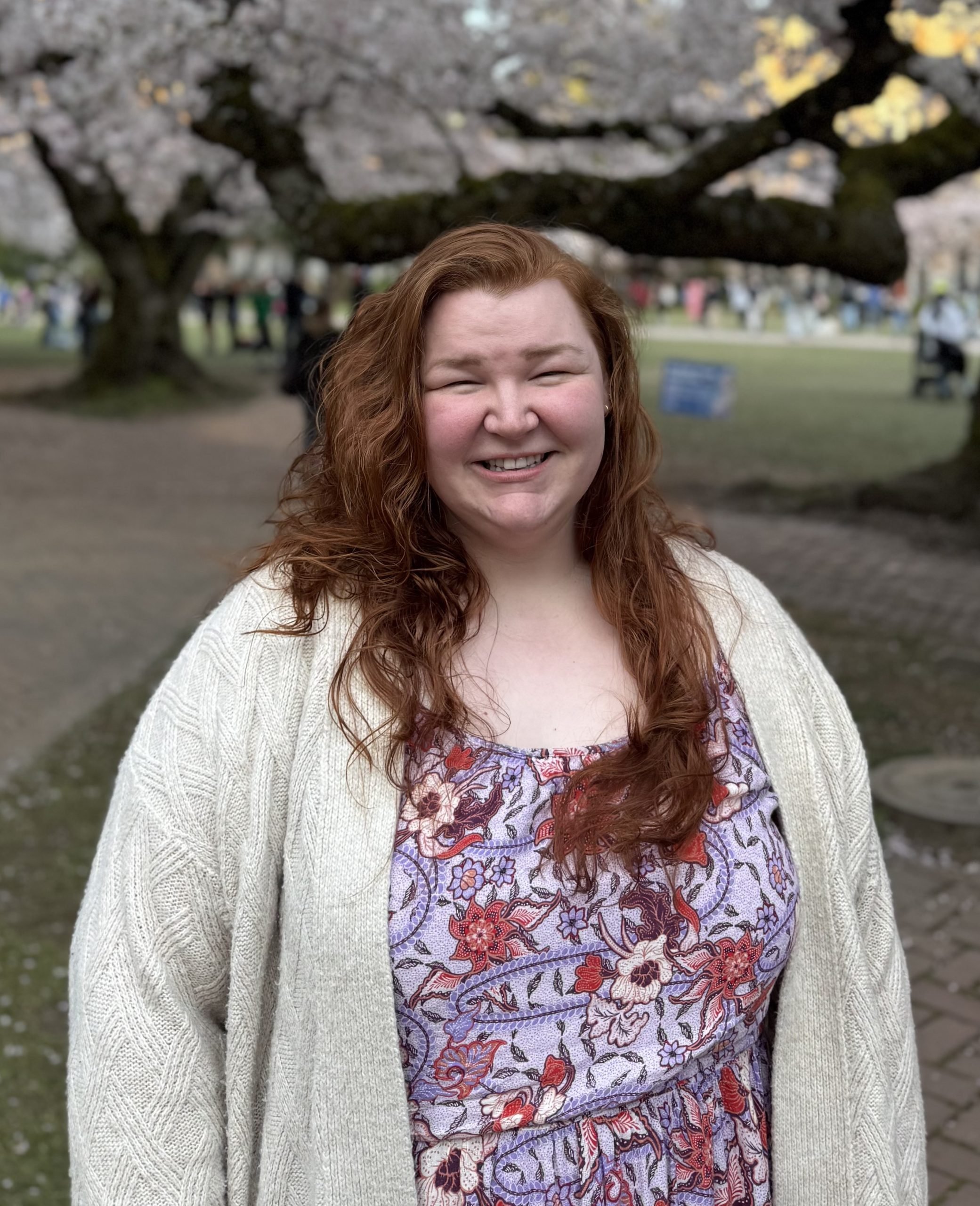 A woman with long curly red hair smiling outdoors in front of a blossoming tree, wearing a patterned dress and a light-colored cardigan.