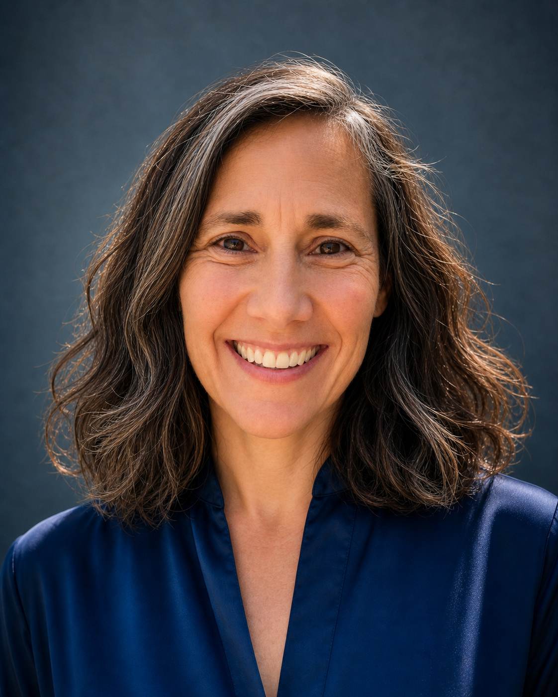 A smiling woman with shoulder-length wavy brown hair, wearing a navy blue satin blouse, standing against a dark blue background.