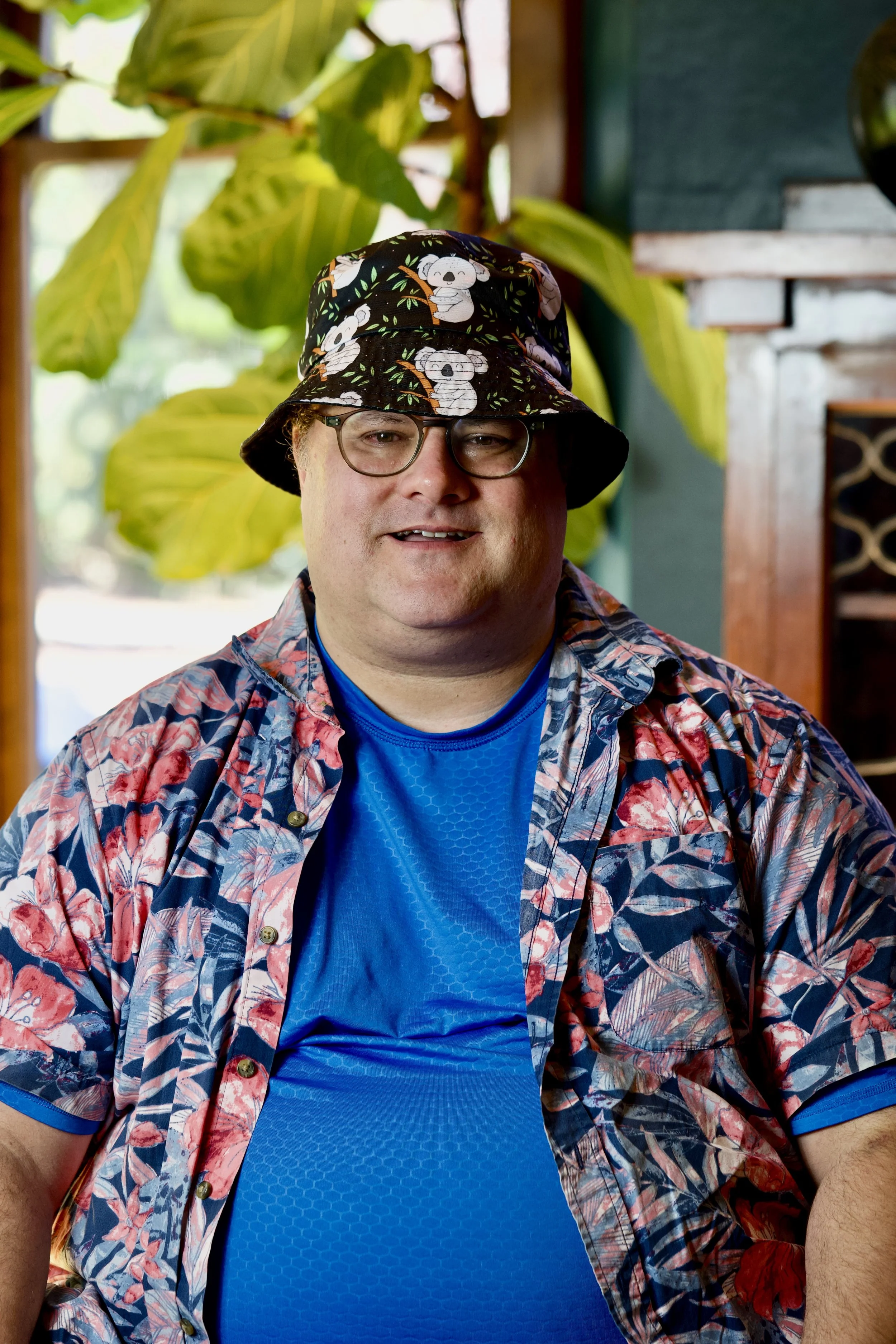 A man wearing glasses, a colorful floral shirt, a blue undershirt, and a bucket hat with koala bear prints sitting in front of large green leaves and a wooden cabinet.