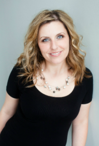 Portrait of a smiling young woman with wavy blonde hair, wearing a black top and a statement necklace, against a light-colored background.