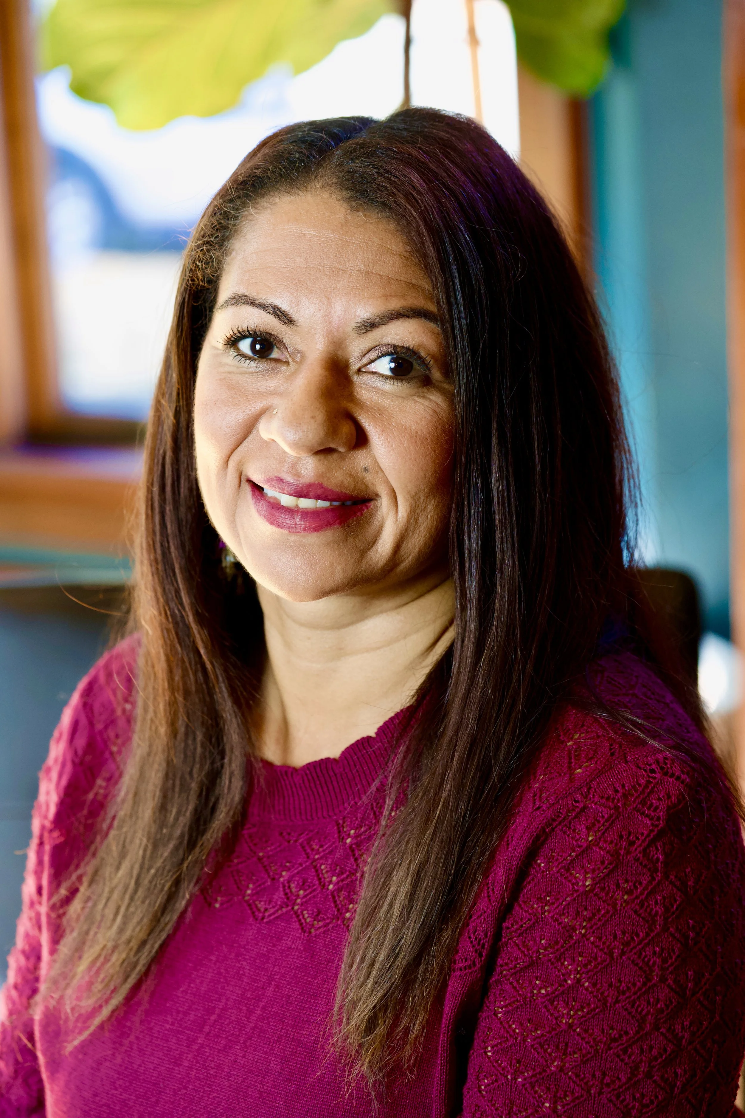 Close-up of a smiling woman with long, straight brown hair wearing a maroon sweater, indoors near a window with natural light.