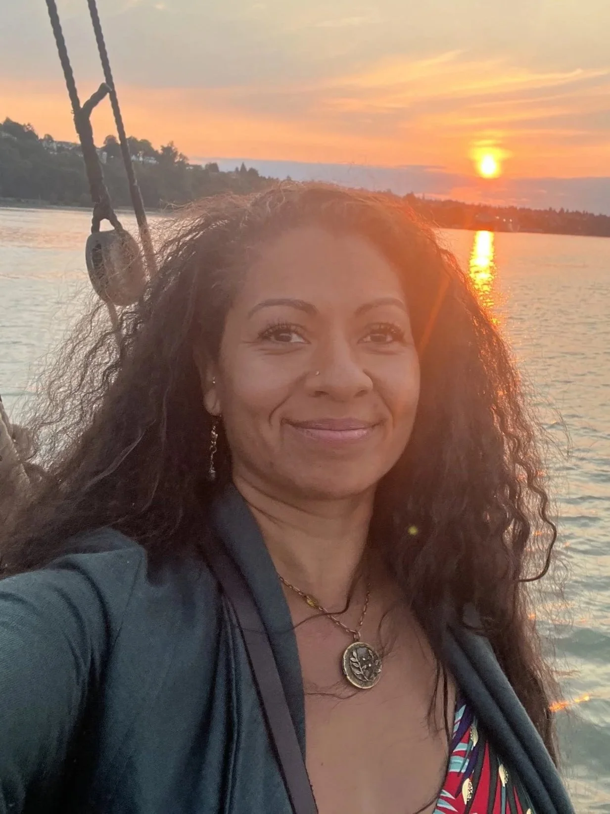 A woman with curly hair smiling at sunset on a boat, with the water and a distant shoreline in the background.