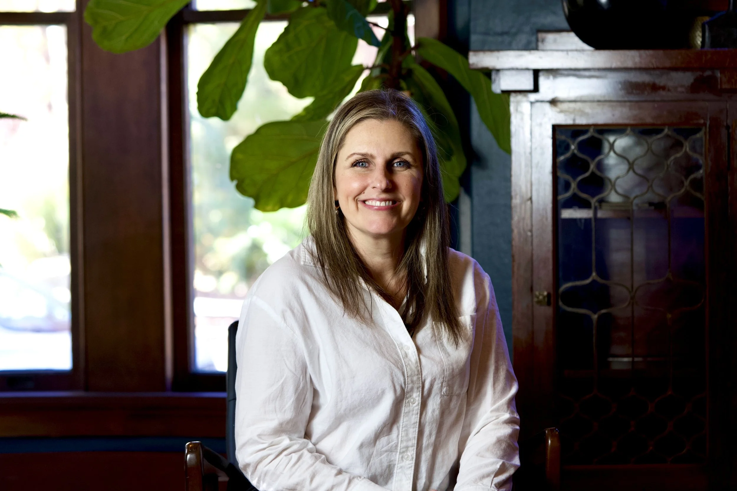 A woman with long blonde hair and blue eyes smiling, sitting inside near a window with large green leaves behind her, wearing a white shirt.