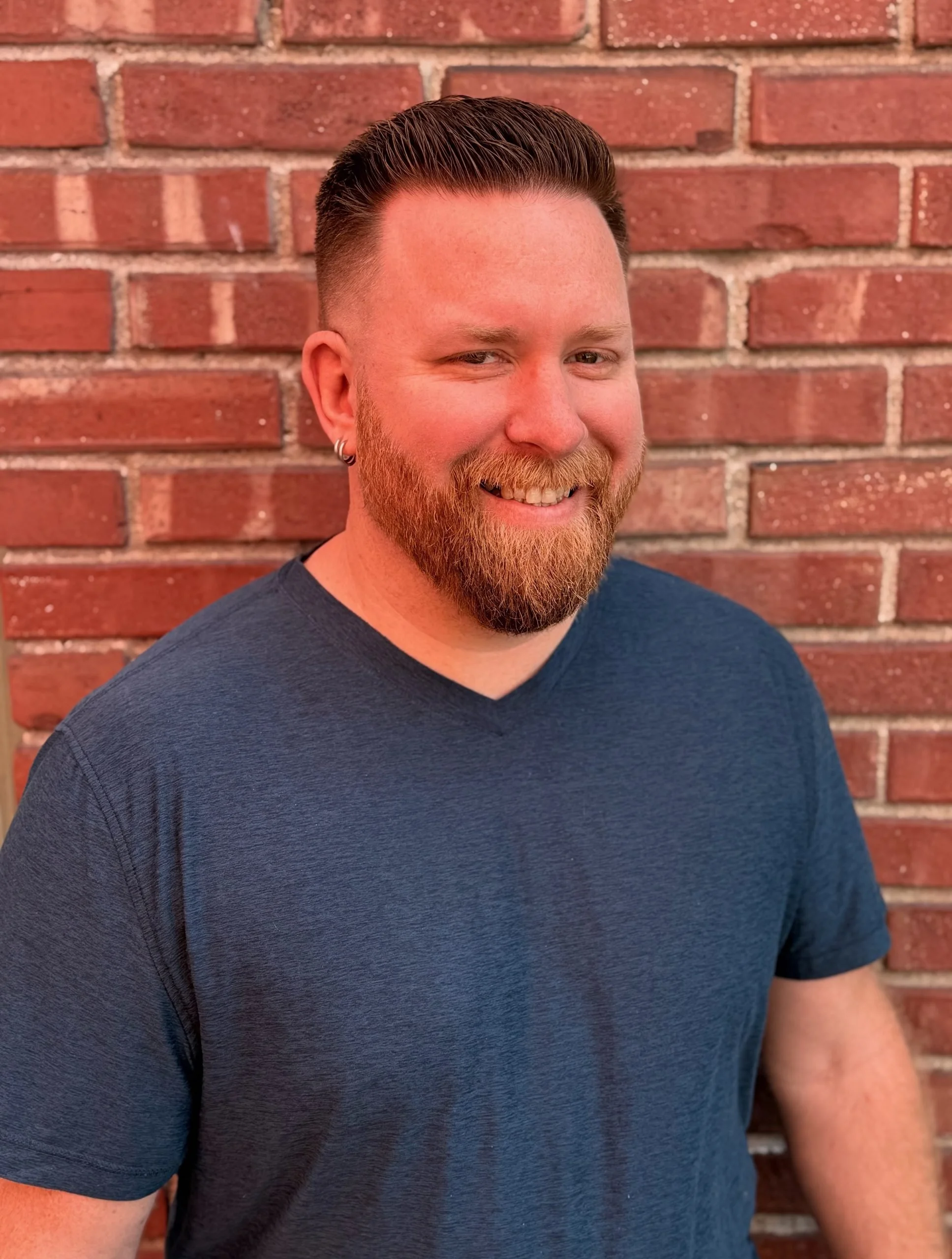 A smiling man with a red beard, wearing a navy blue t-shirt, standing in front of a red brick wall.