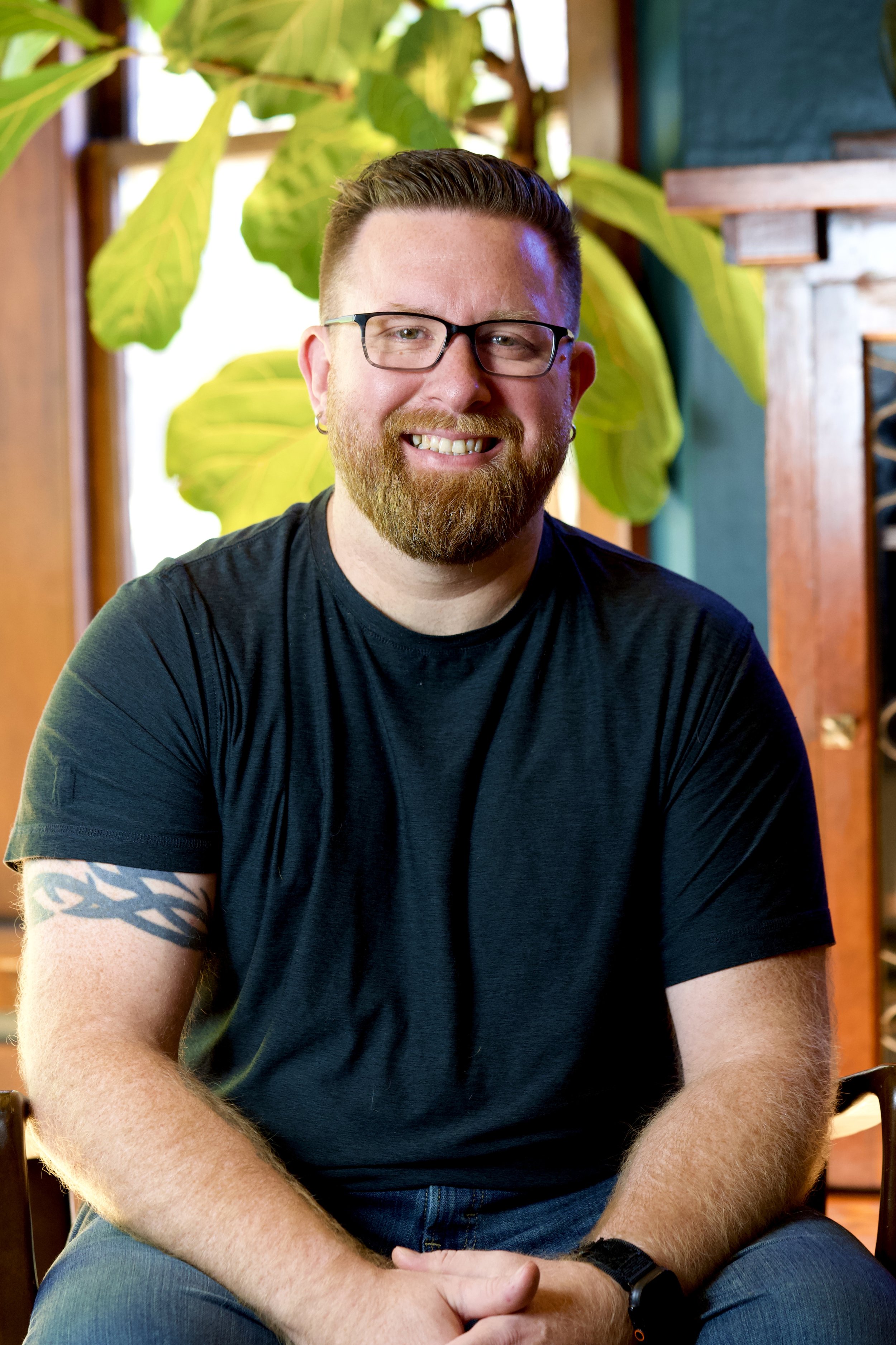 A smiling man with glasses and a beard, sitting indoors with a large green plant and wooden furniture in the background.