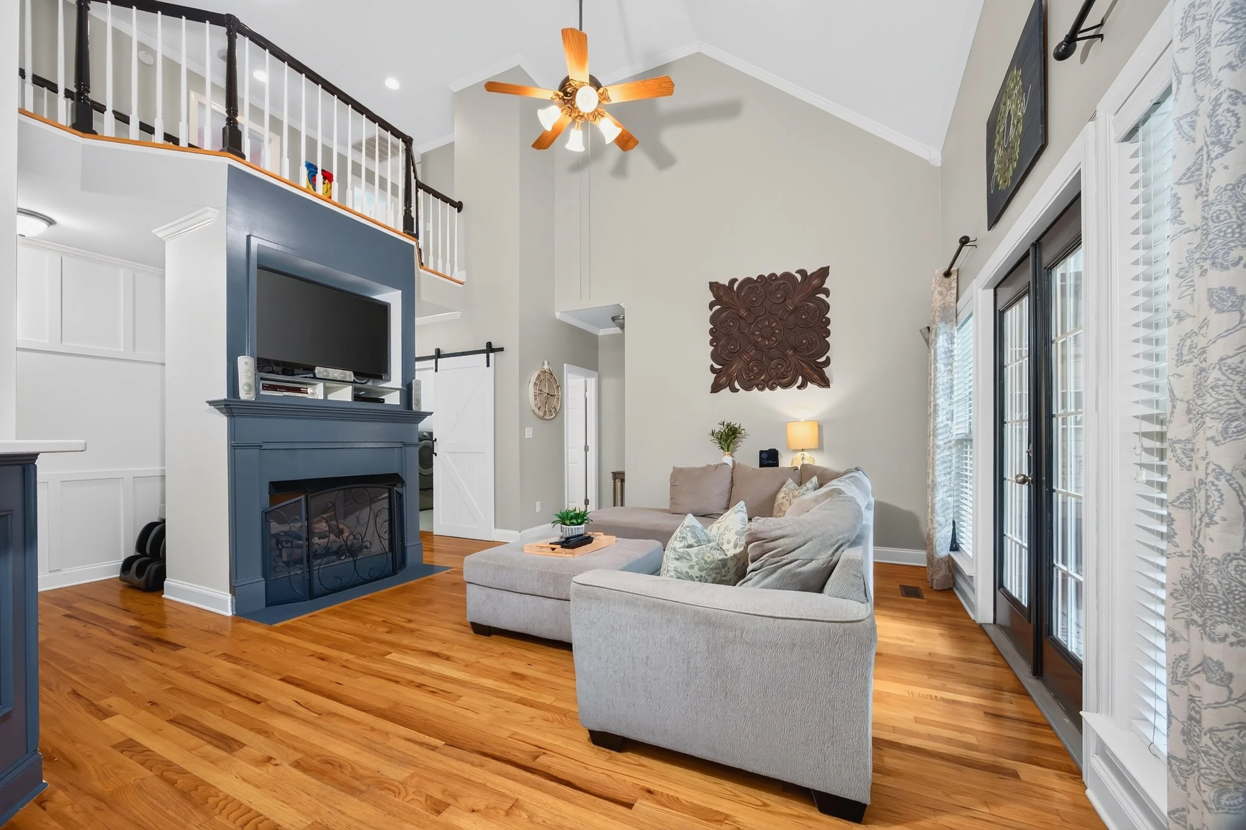 A living room with a high ceiling, ceiling fan, wall art, large windows with curtains, a beige sectional sofa with decorative pillows, a coffee table, and a fireplace with built-in TV area. Hardwood floors and a decorative clock on the wall are also 