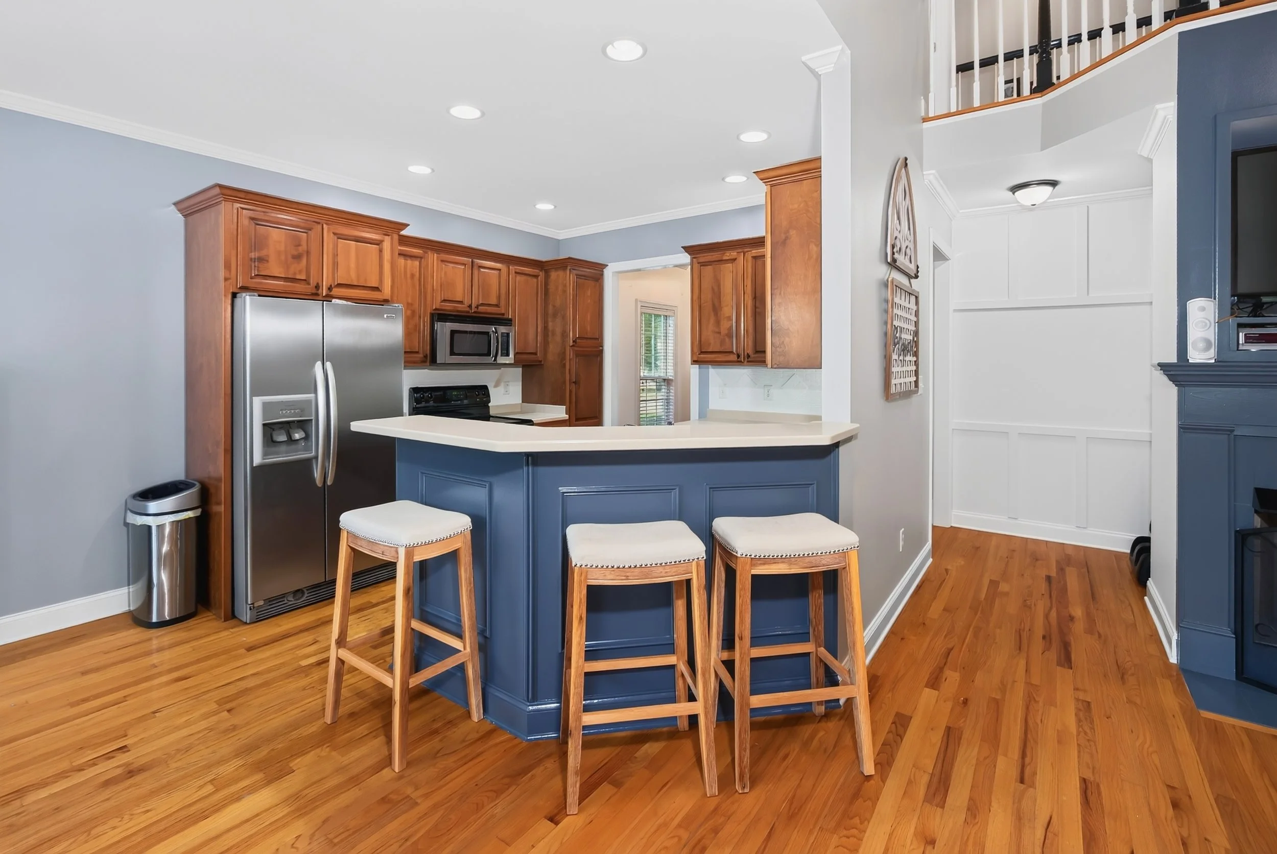 Kitchen with wooden cabinets, stainless steel refrigerator, microwave, and a blue kitchen island with three bar stools, hardwood floors, and a staircase in the background.
