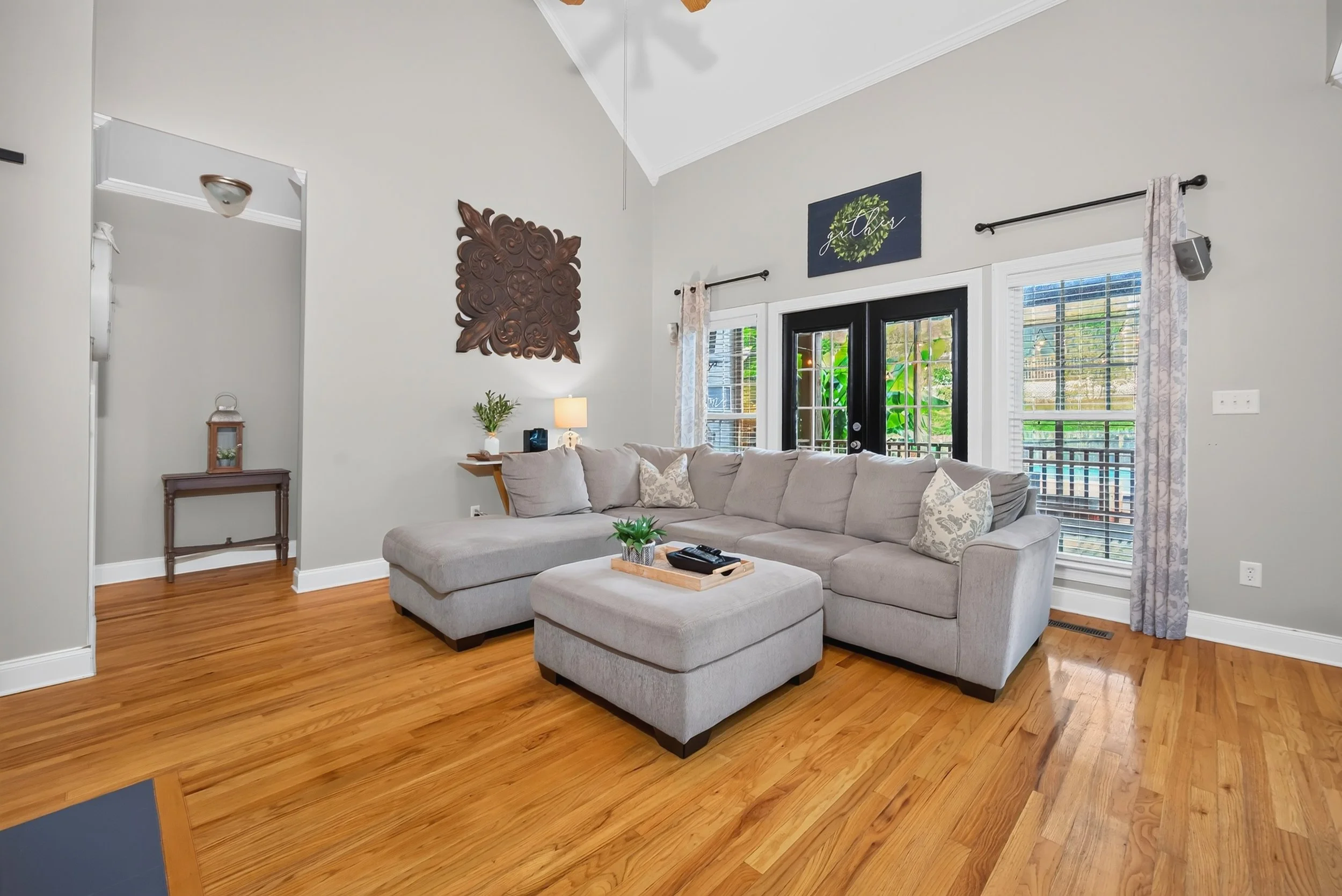 Living room with beige sectional sofa, wooden flooring, and large windows with white curtains. Decor includes wall art, a small table with a lamp, and outdoor greenery visible through the windows.