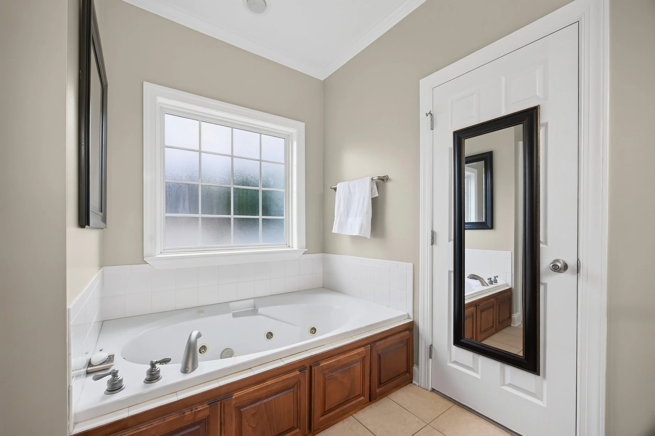 Bathroom with a large window, a bathtub with jets, a wooden cabinet underneath, and a tall mirror on the door.
