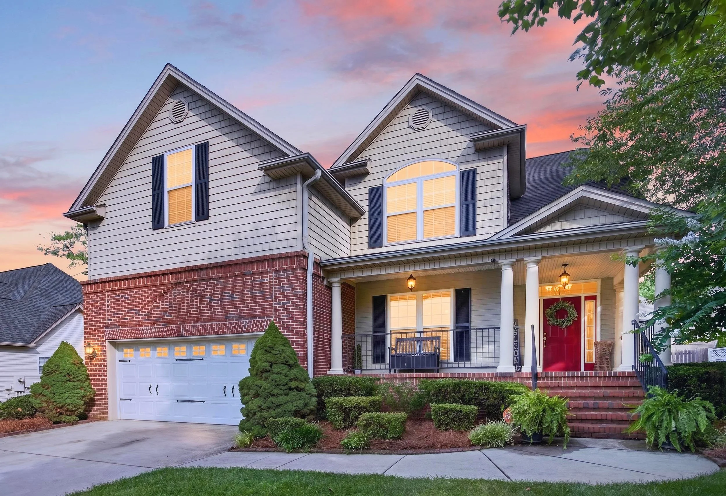 Front view of a two-story suburban house with a brick lower level and beige siding upper level, white garage door, and red front door, with a well-landscaped yard and trees, during sunset.