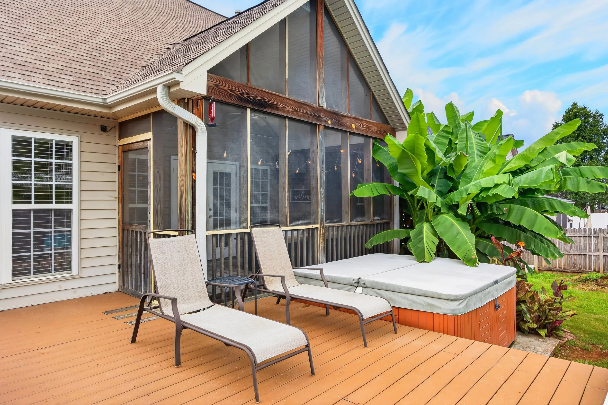 Backyard patio with wooden deck, lounge chairs, a large green banana plant, and a screened-in porch area attached to a house with beige siding and a brown shingle roof.