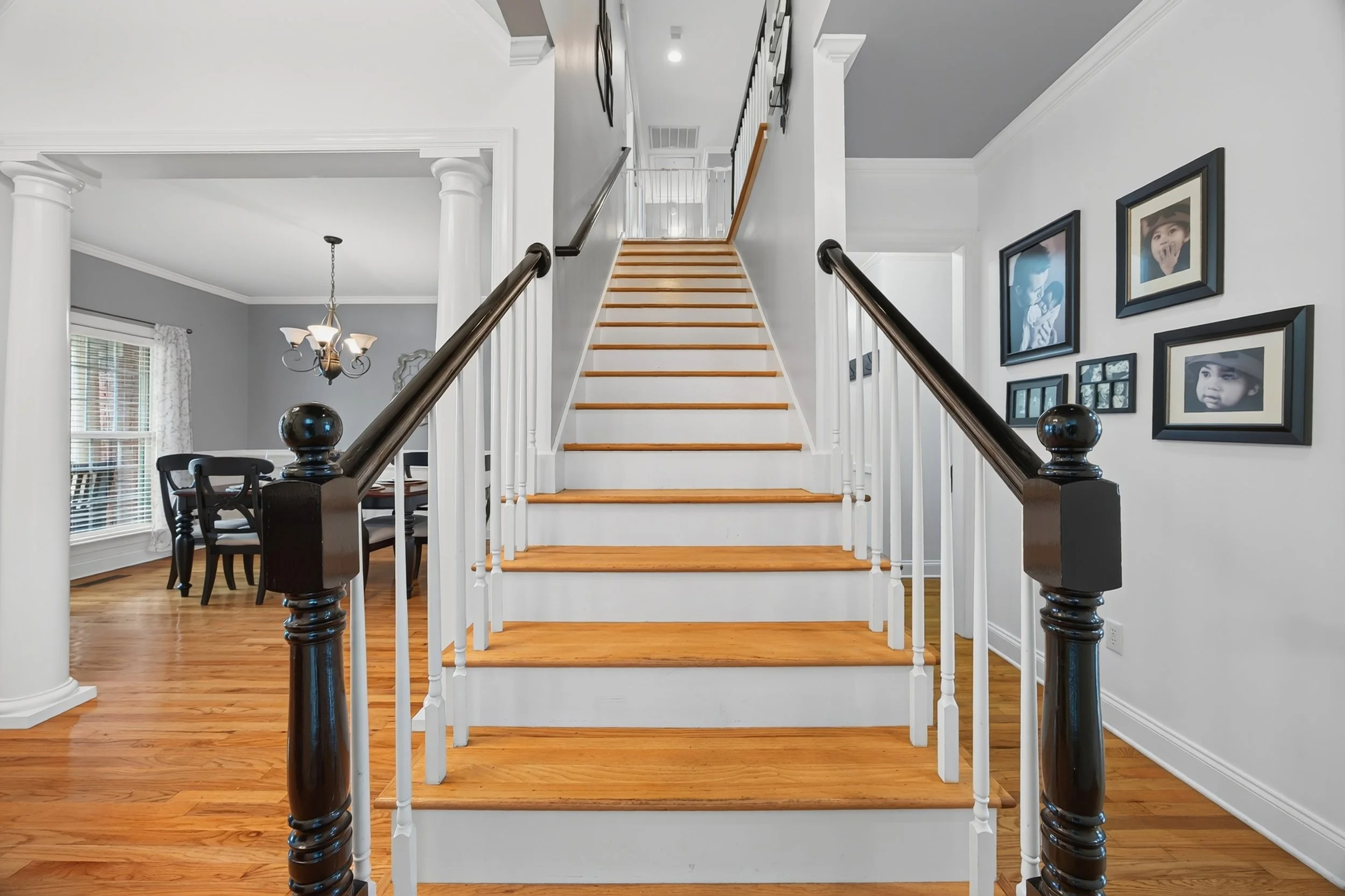 Interior view of a home staircase with wooden steps, white risers, and black handrails on both sides. To the left, there is a dining area with a black dining table and chairs underneath a chandelier, next to a window with white blinds and curtains. F