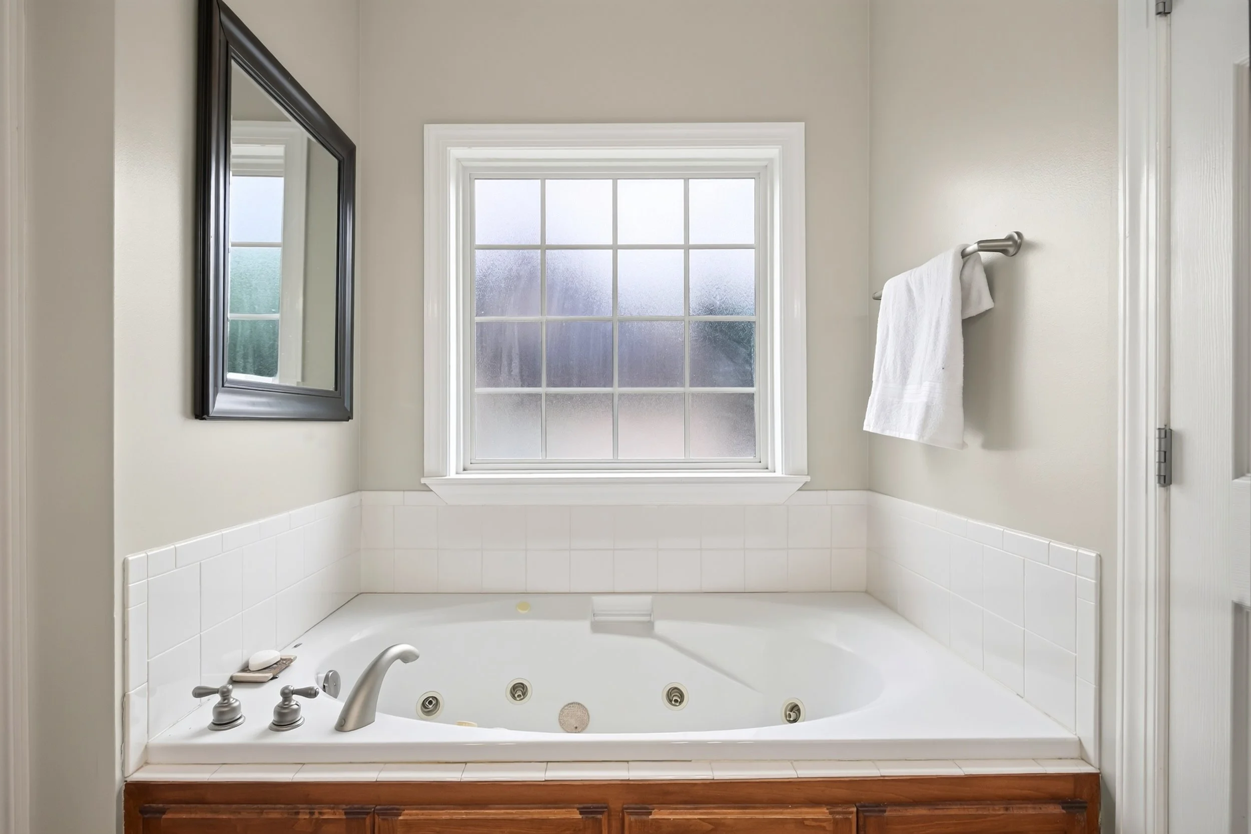 A bathroom with a large frosted window above a white bathtub, a wall-mounted mirror to the left, and a white towel hanging on a silver towel bar to the right.
