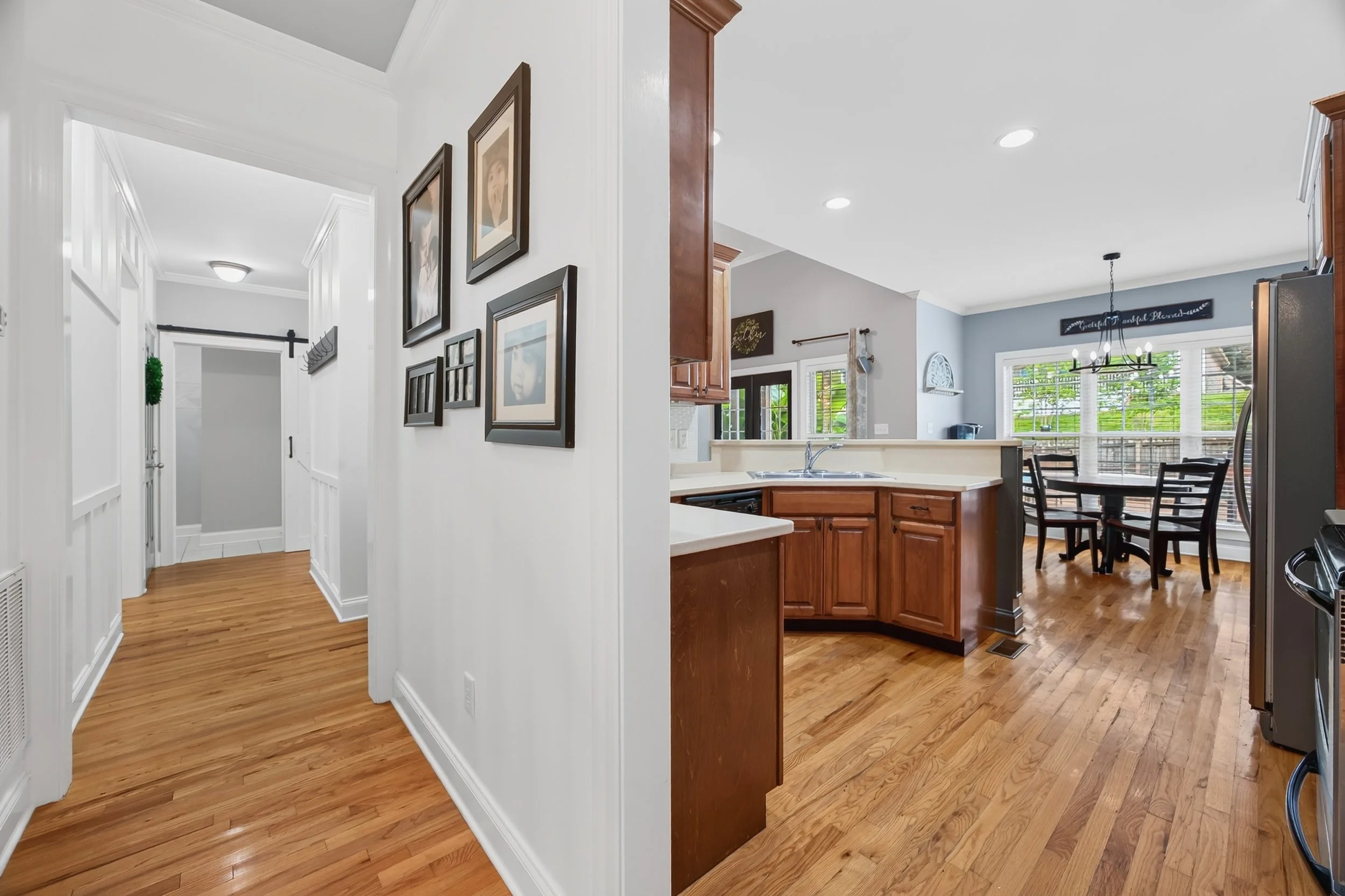 Interior view of a house showing a hallway with wooden floors, white walls, framed pictures, and a glimpse of the kitchen and dining area with large windows and a chandelier.