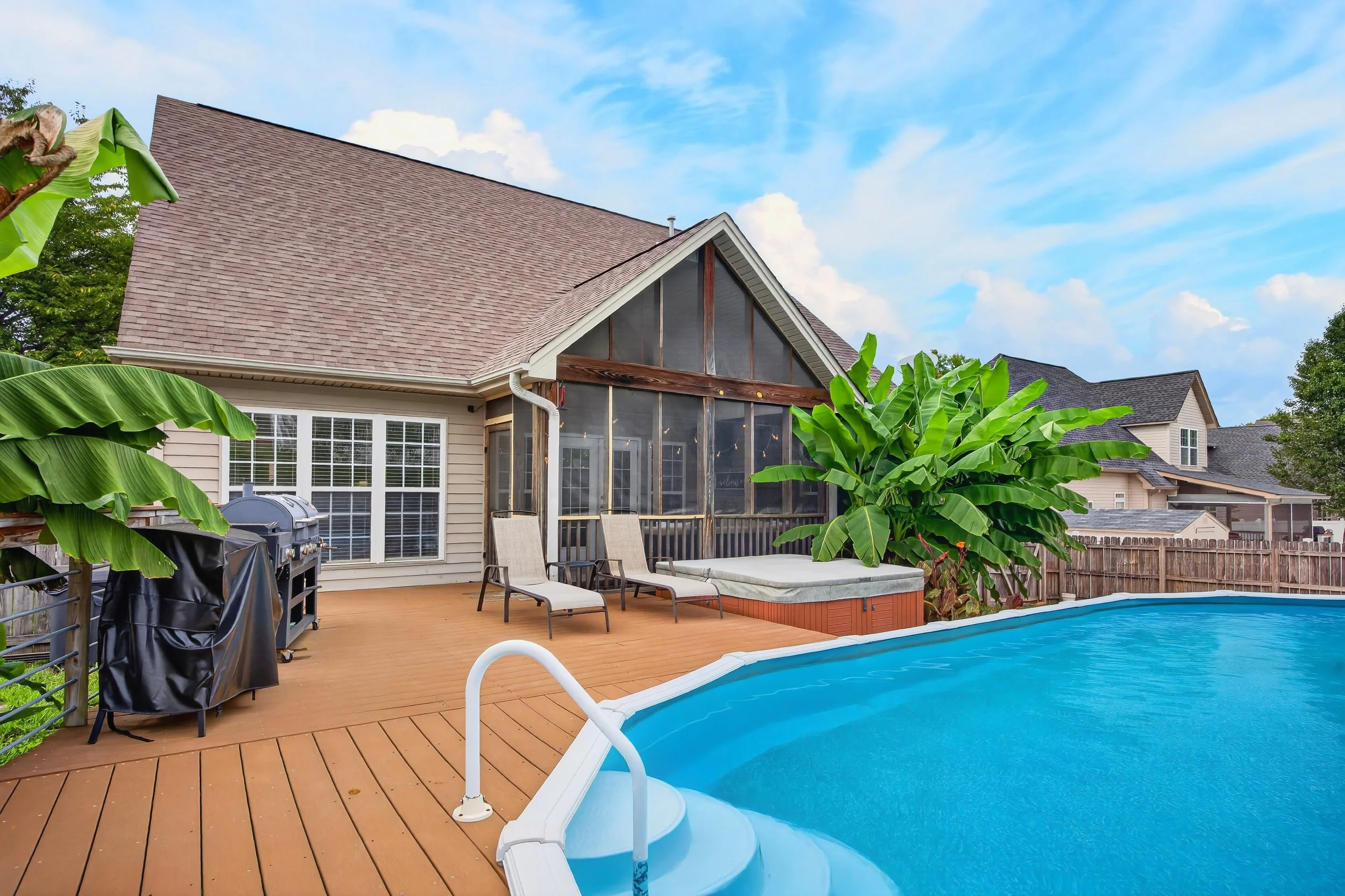 Backyard with swimming pool, lounge chairs, barbecue grill, tropical plants, house with screened porch, and a blue sky with some clouds.