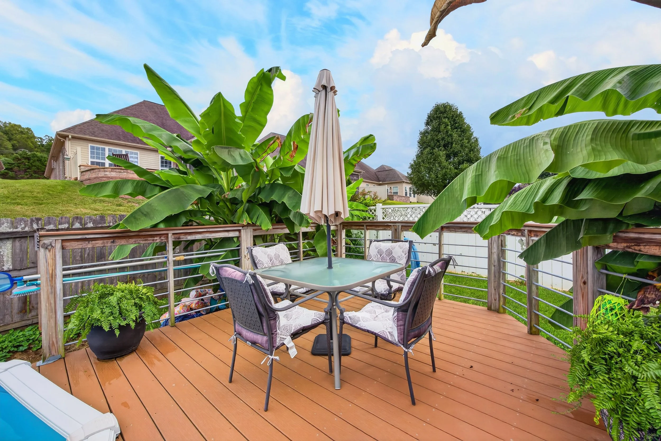 A backyard patio with wooden deck, surrounded by large green banana leaves and ferns, featuring a glass outdoor table with six chairs, a closed beige umbrella, and a view of neighboring houses and trees.