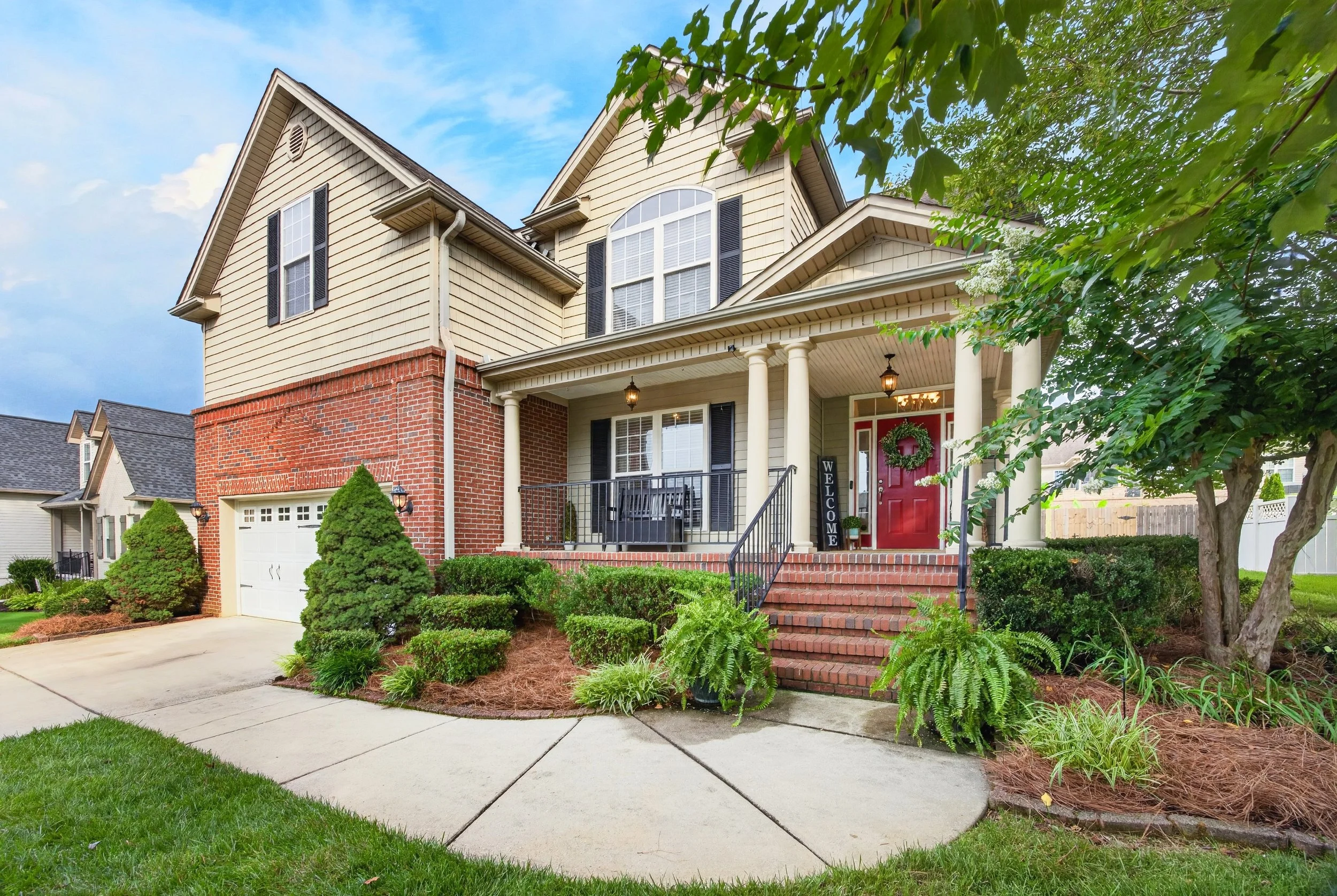 Front view of a two-story house with a red front door, brick and siding exterior, white columns, and a front porch. The yard has green shrubs, trees, and a concrete walkway leading to the stairs.