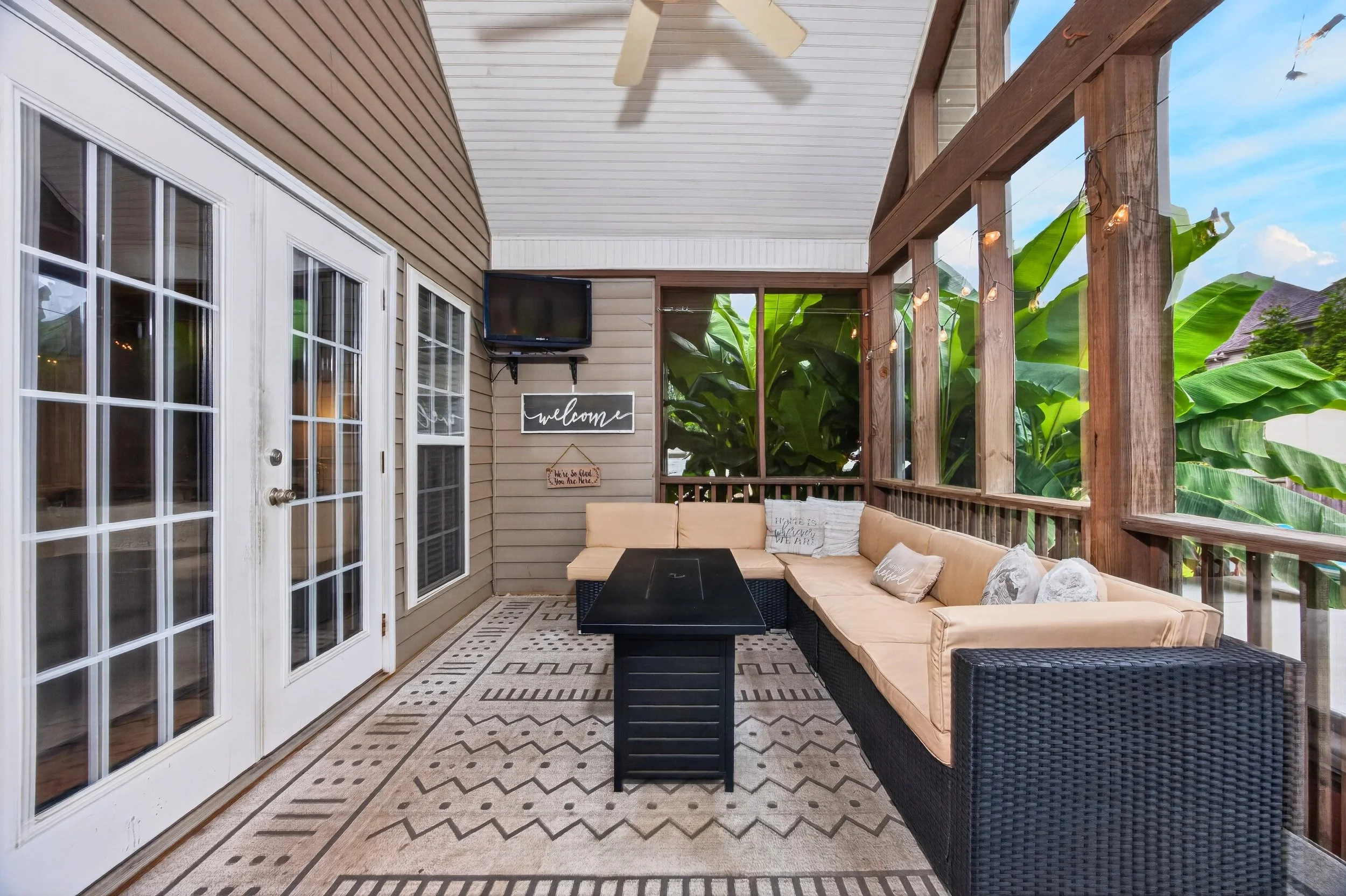 Covered outdoor patio with beige sectional sofa, black wicker bases, black coffee table, patterned outdoor rug, string lights, ceiling fan, wall-mounted TV, welcome signs, white French doors, large green leaves outside, blue sky with clouds.