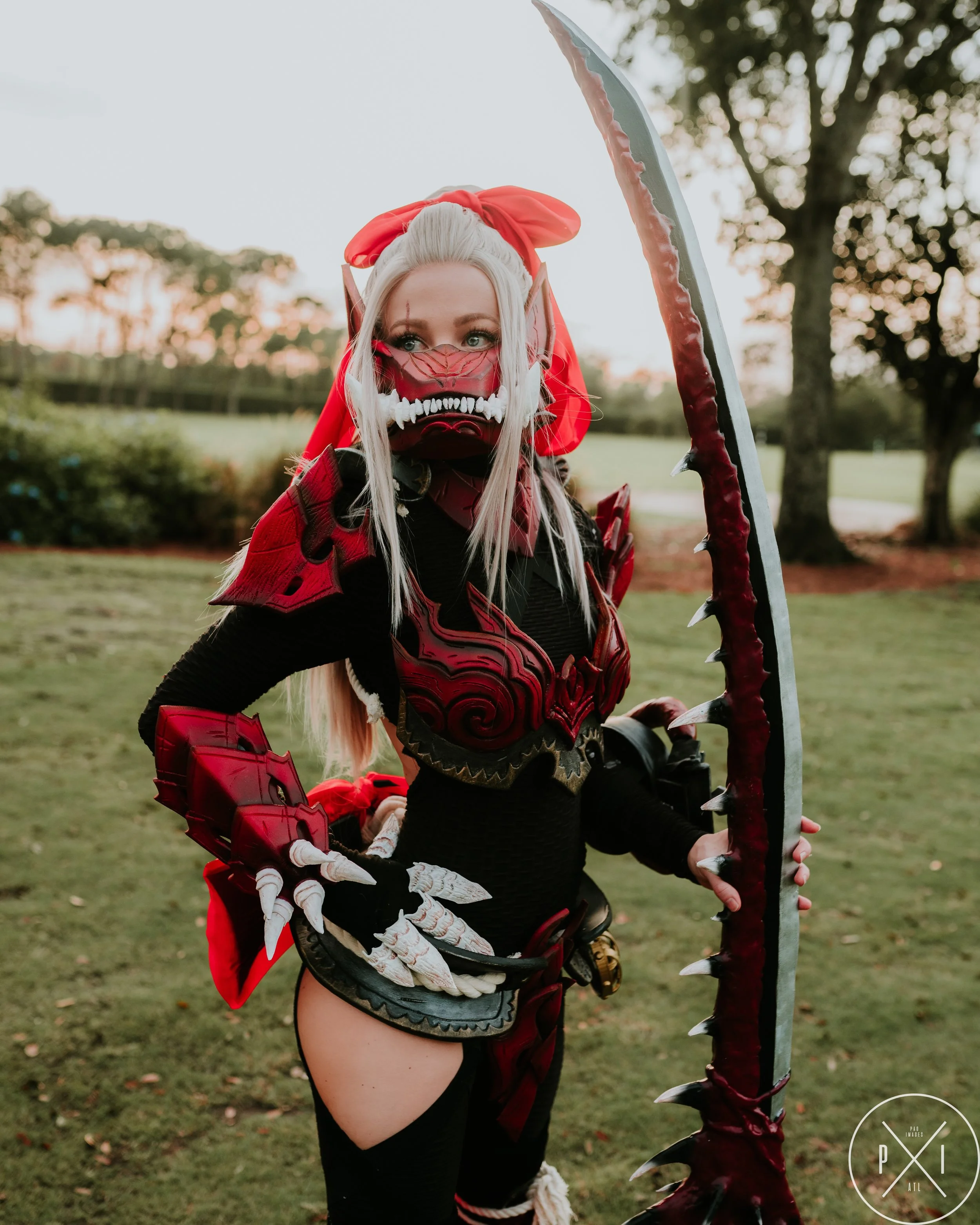 Person dressed in a detailed red and black fantasy armor costume holding a large spiked weapon, outdoors on grass with trees in the background.