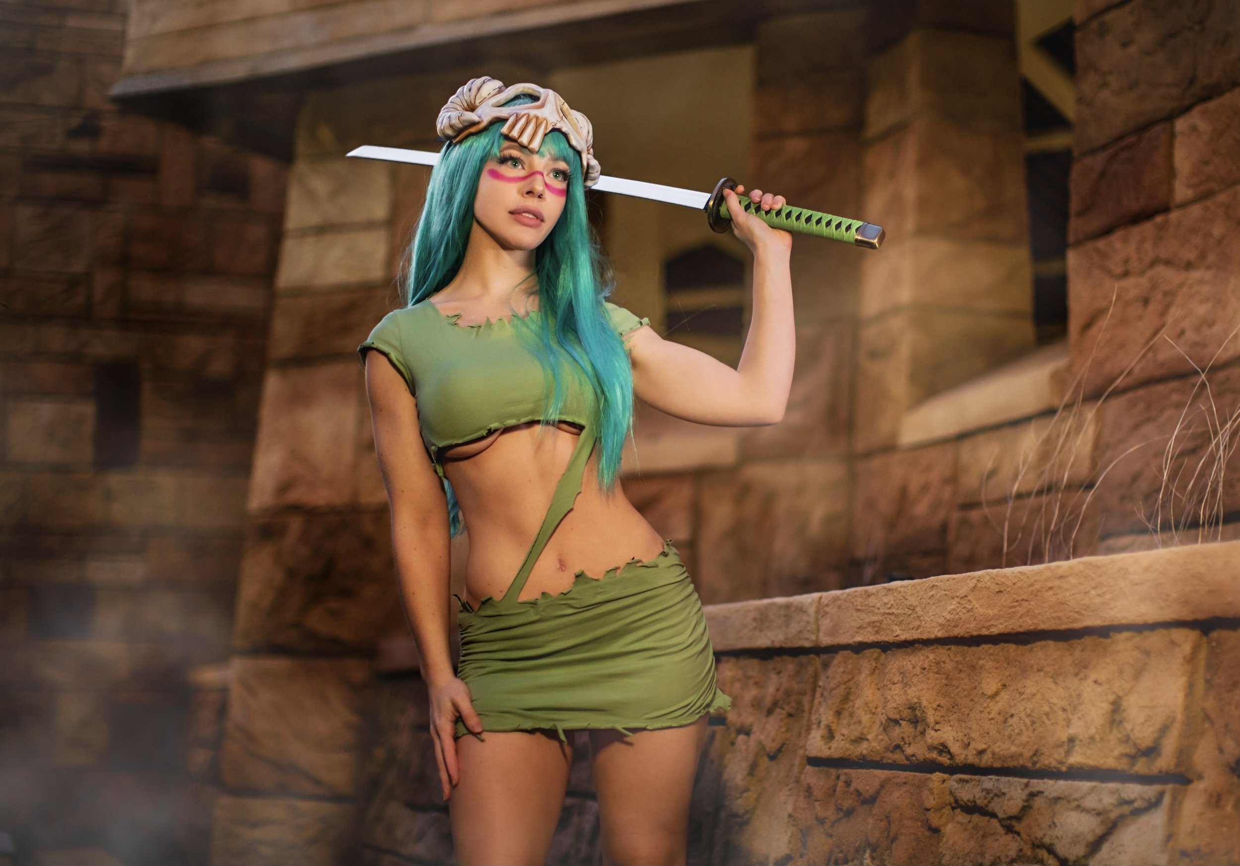 A woman with long teal hair and pink face paint holding a sword on her shoulder, wearing a green shredded outfit in a rocky, desert-like environment.