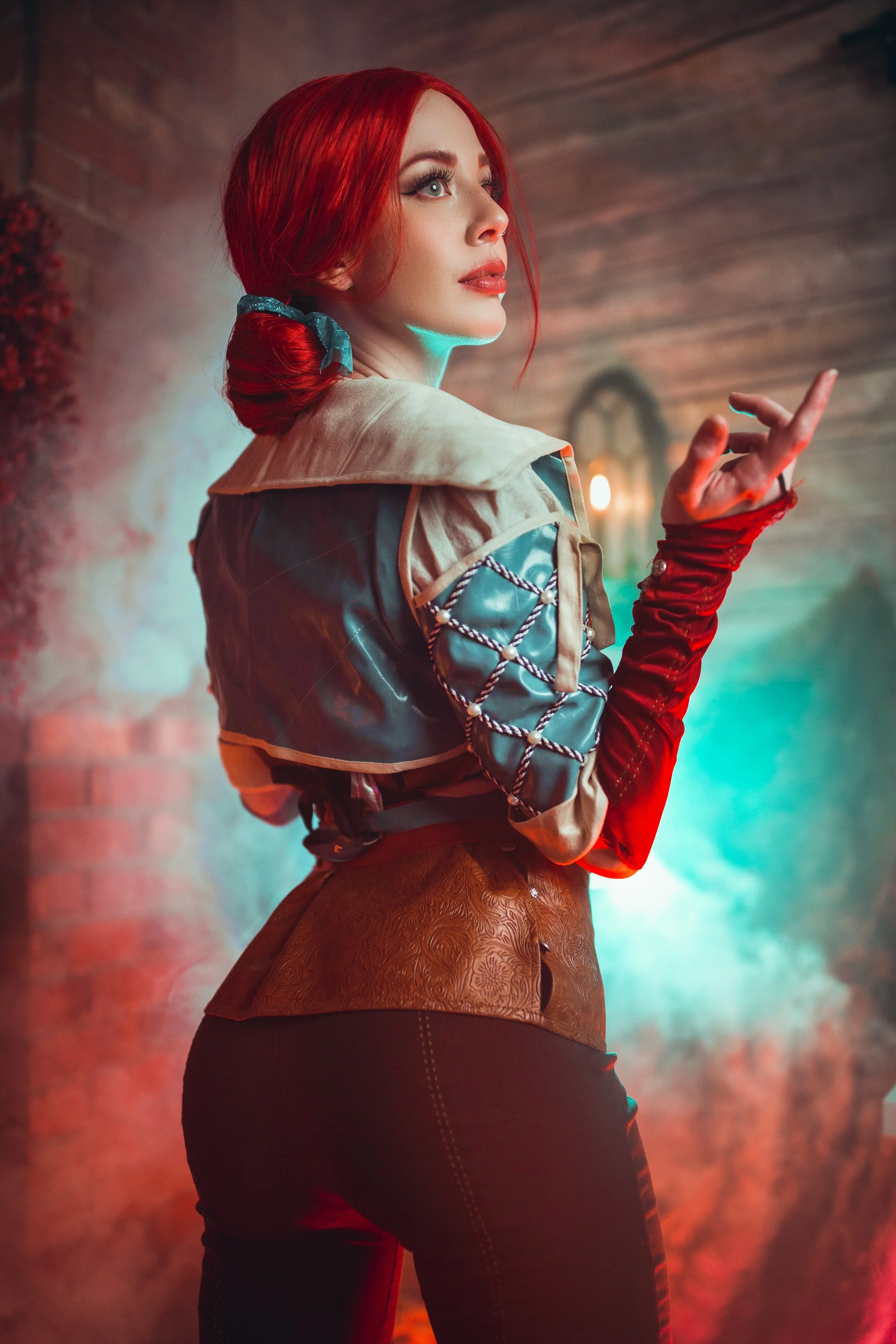 A woman with red hair styled in a low bun tied with a blue ribbon, wearing a medieval or fantasy-inspired costume with a beige and blue top, standing against a brick wall in a smoky, colorful atmosphere with red, blue, and orange lighting.