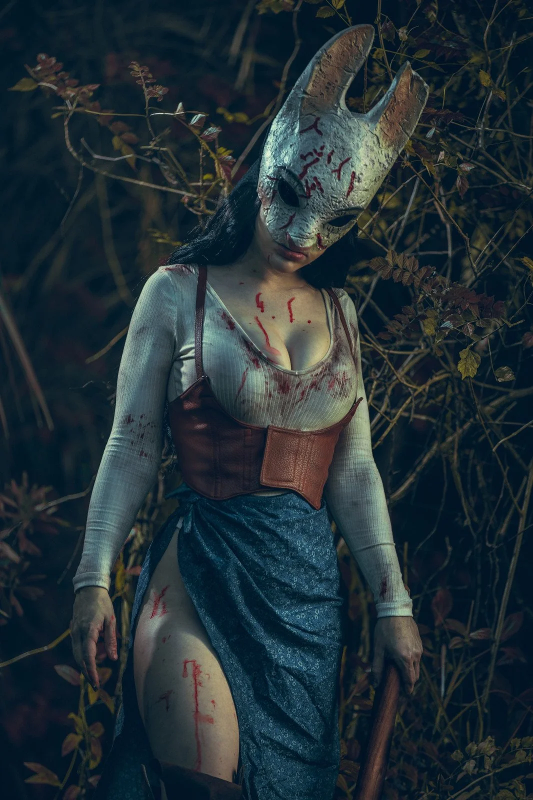 A person wearing a creepy white rabbit mask with black eyes, standing outdoors among branches and leaves. The person is dressed in a bloodstained beige top, a brown corset, and a blue skirt with a slit. The scene appears dark and eerie.