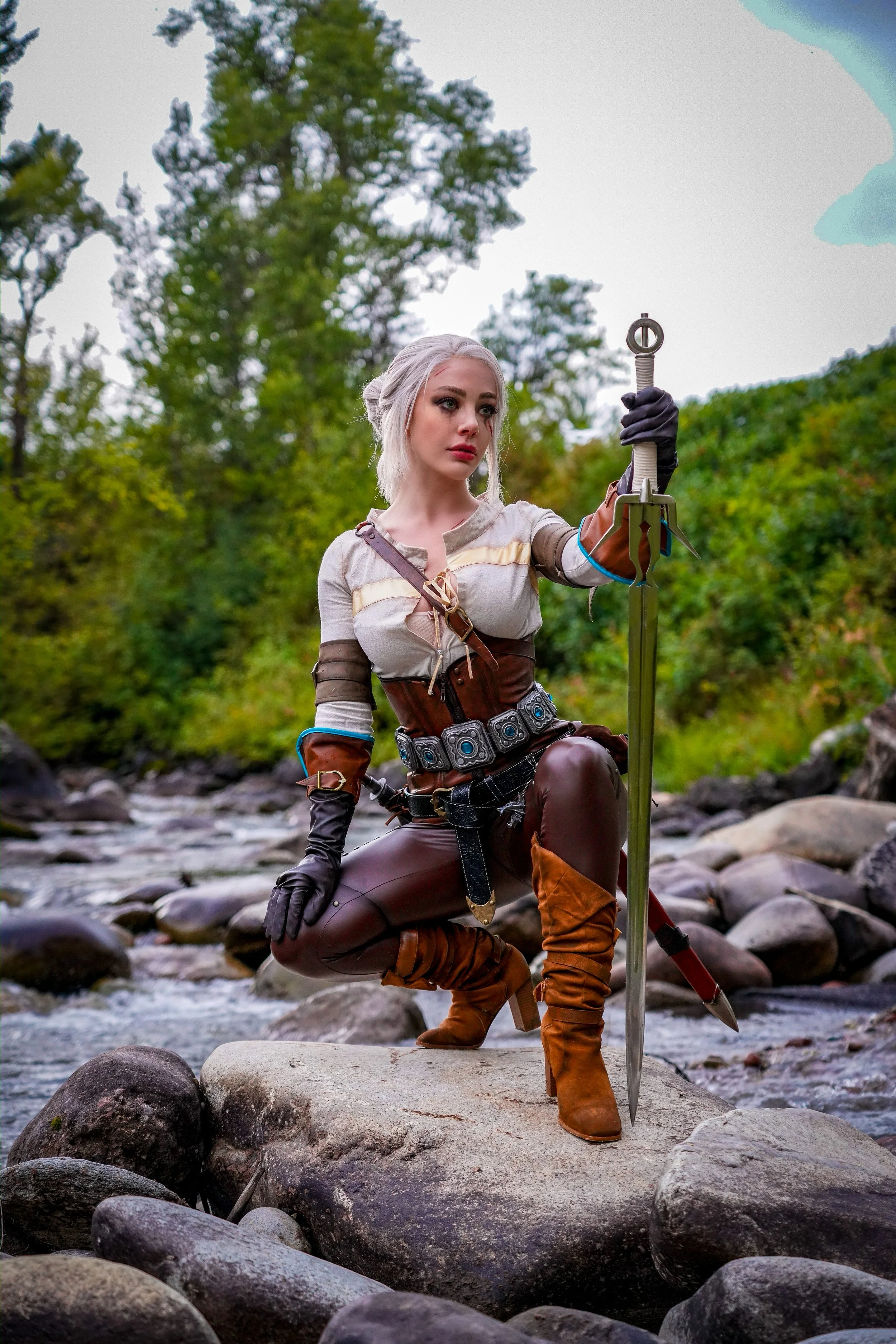 A woman dressed as a fantasy warrior, kneeling on rocks in a river, holding a sword, with a forest in the background.