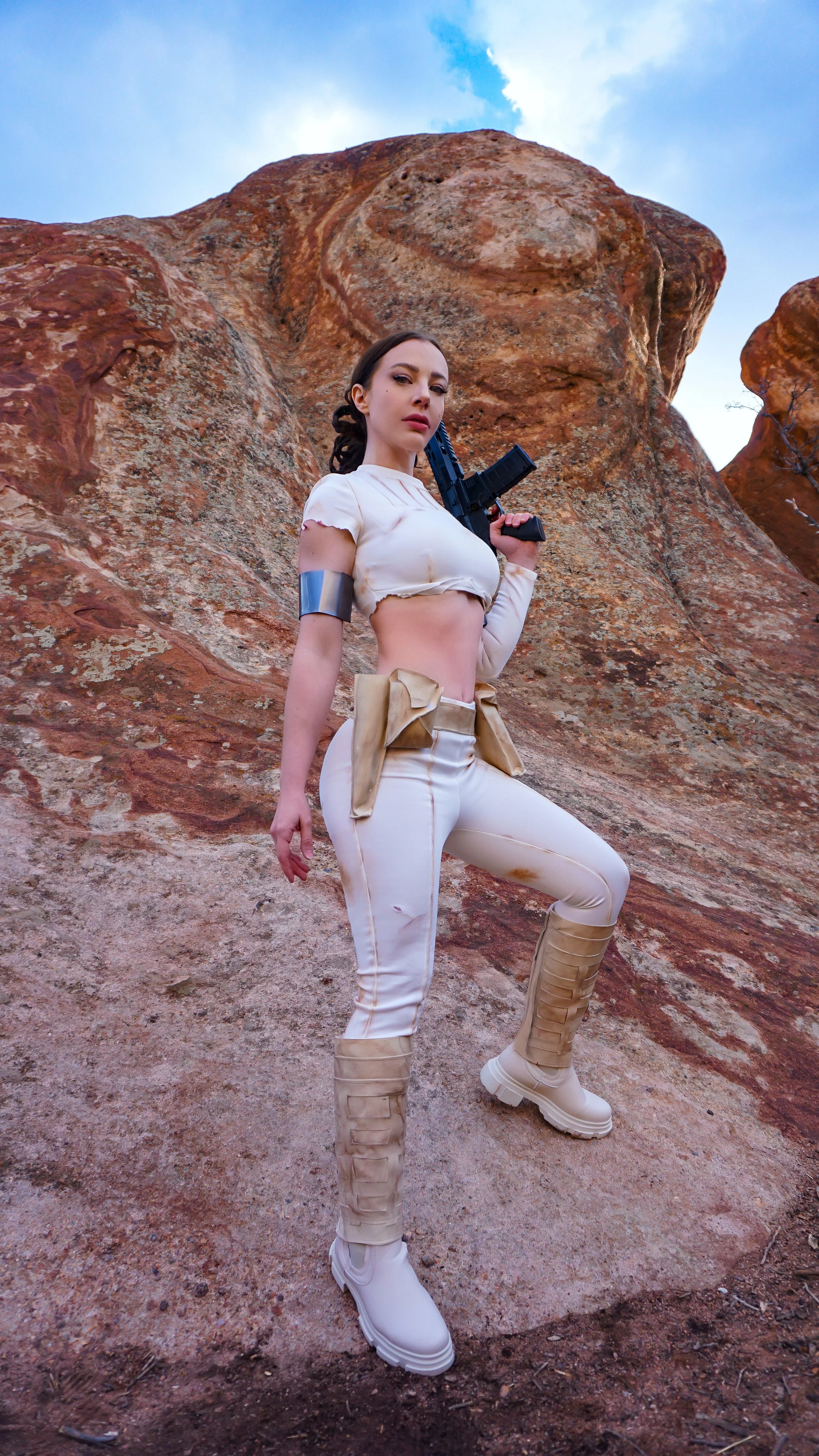 A woman dressed in a futuristic, beige and white outfit with combat boots, holding a black rifle, standing in a rocky, desert landscape with a blue sky above.