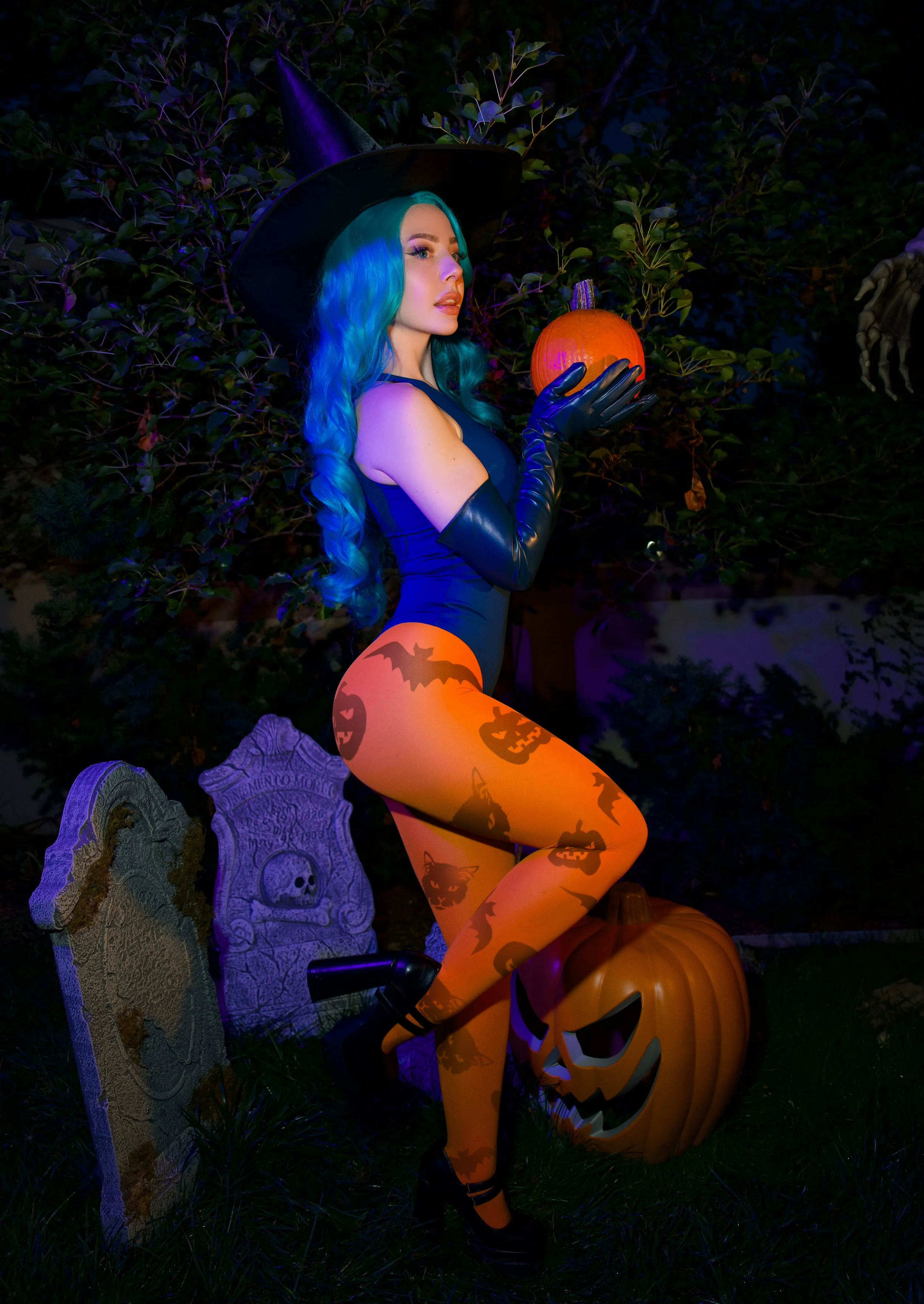 A woman dressed as a witch, with blue and green hair and wearing a black hat, holds a small pumpkin in a dark outdoor setting decorated for Halloween with tombstone and pumpkin decorations.
