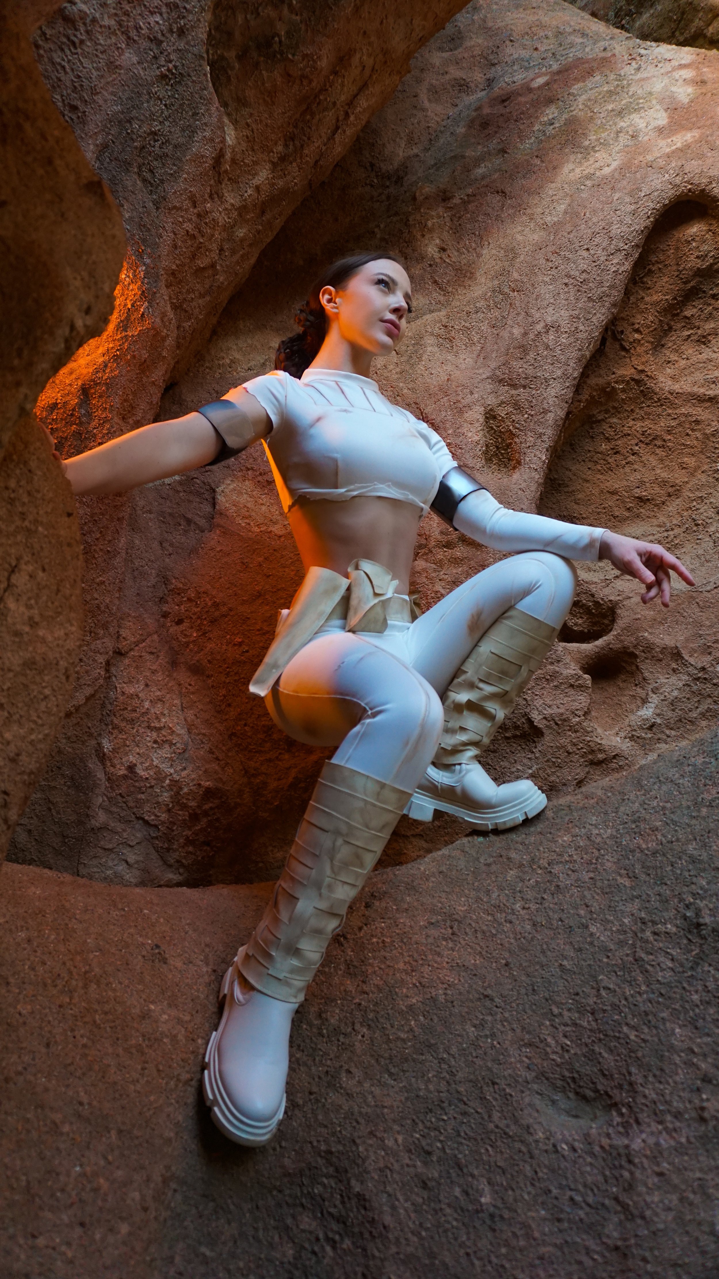 A woman dressed in a futuristic white space suit with beige accents poses against a textured rock wall, with her arms outstretched and one knee raised.