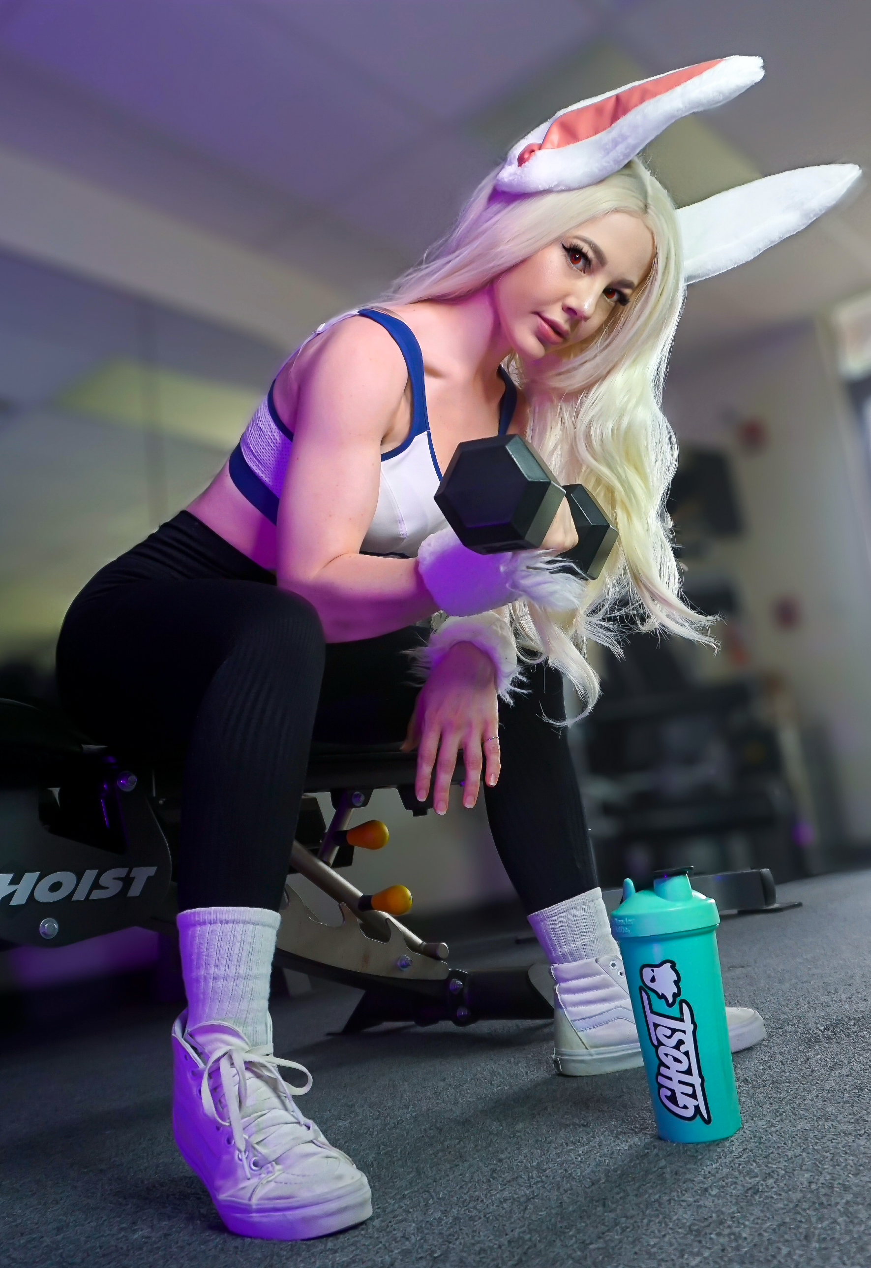 A woman dressed as a bunny with long white hair, bunny ears, and furry gloves, lifting a dumbbell in a gym. She is wearing a sports bra, black leggings, white high-top sneakers, and has a blue water bottle with a bunny logo nearby.