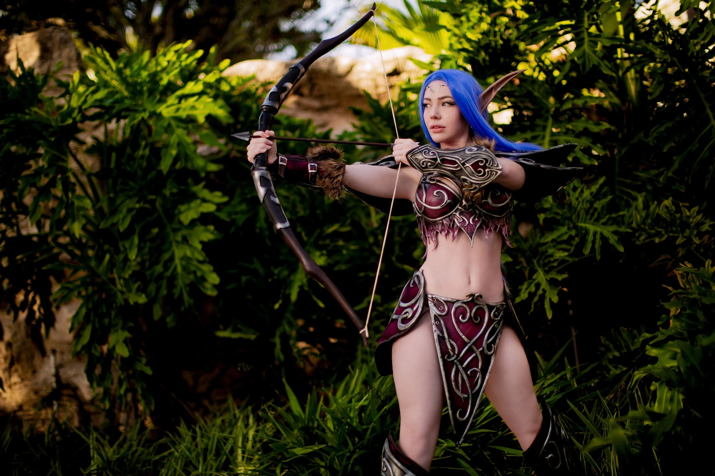 A woman dressed as an elf archer with long blue hair and pointed ears is aiming a bow and arrow outdoors in front of green bushes.