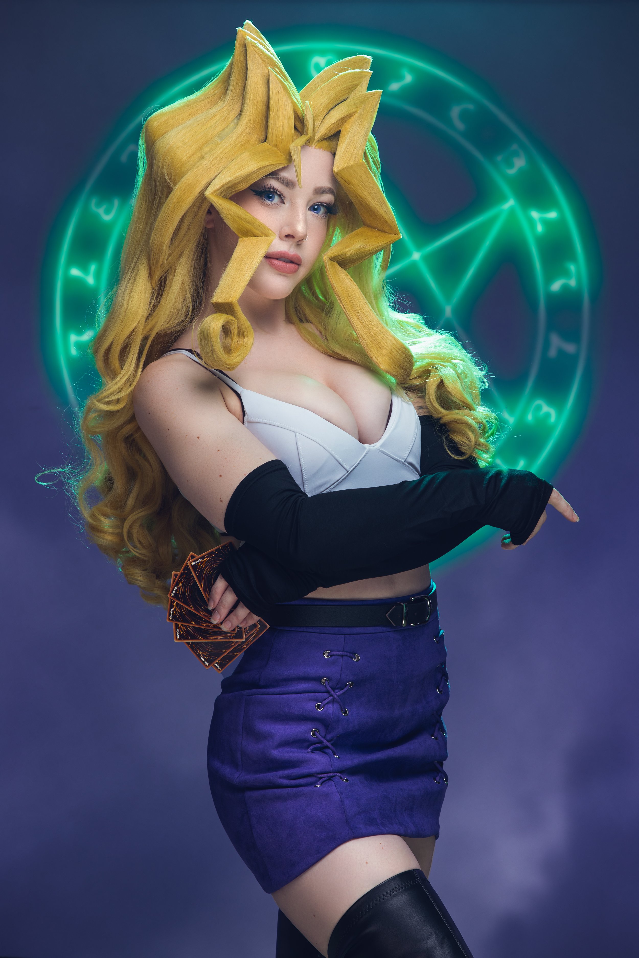 A woman with long, curly, blonde hair wearing a white top with black straps and purple skirt with lace-up details. She holds a fan and is dressed as a character from the anime Yu-Gi-Oh! with stylized blonde hair and a glowing green magic circle behin