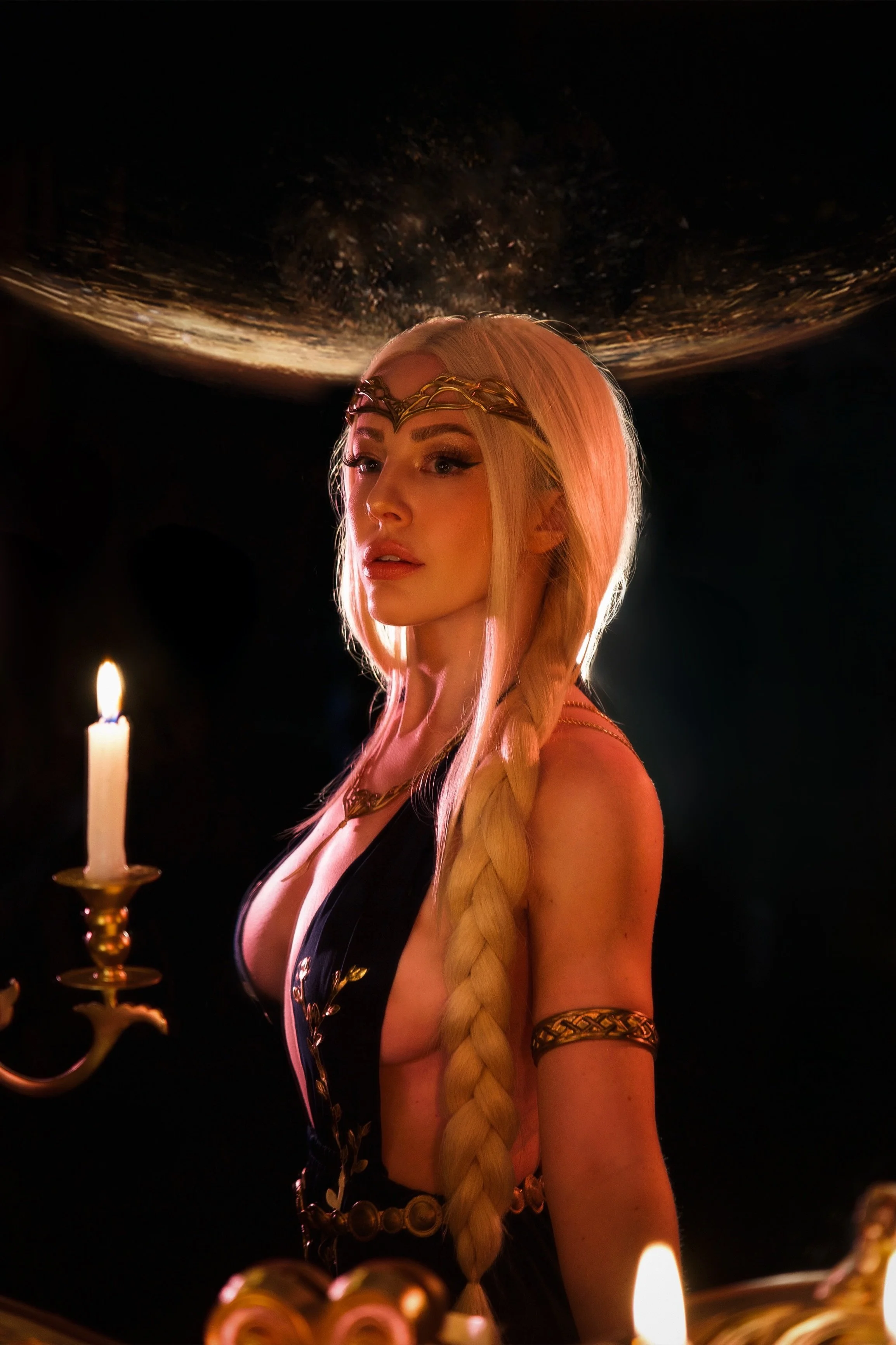 A woman with long blonde braid and a gold headpiece holding a lit candle in a dark setting, with a celestial scene above her.