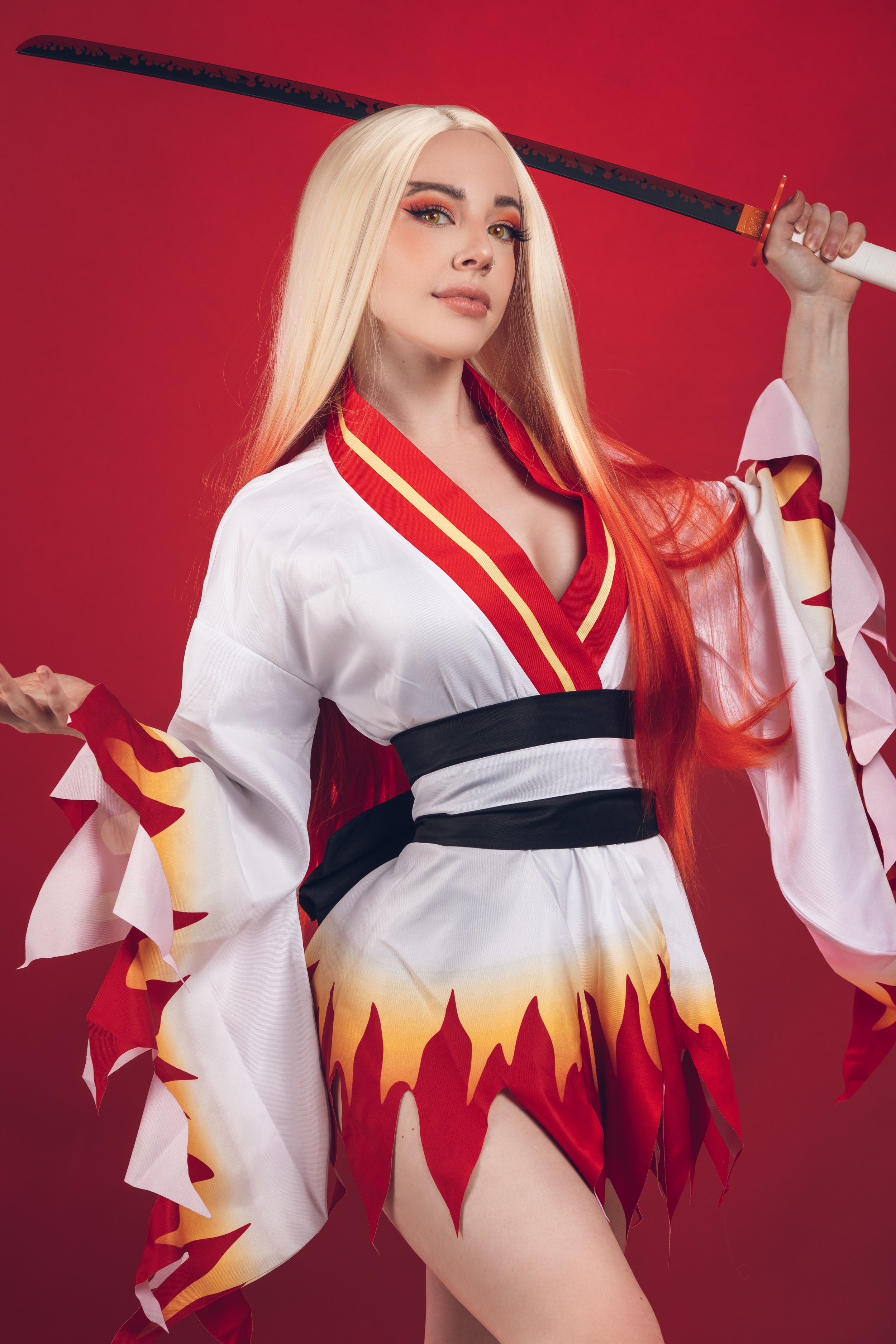 A woman with long blonde and red hair, wearing a traditional red and white Japanese kimono-style outfit with flame patterns, holding a katana sword over her shoulder against a red background.