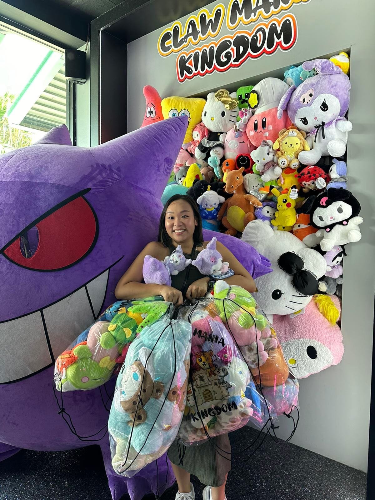 A woman smiling, surrounded by plush toys and colorful bags, in front of a wall labeled 'Claw Mania Kingdom' featuring a large plush of a purple cartoon character and a wall full of various stuffed animals.
