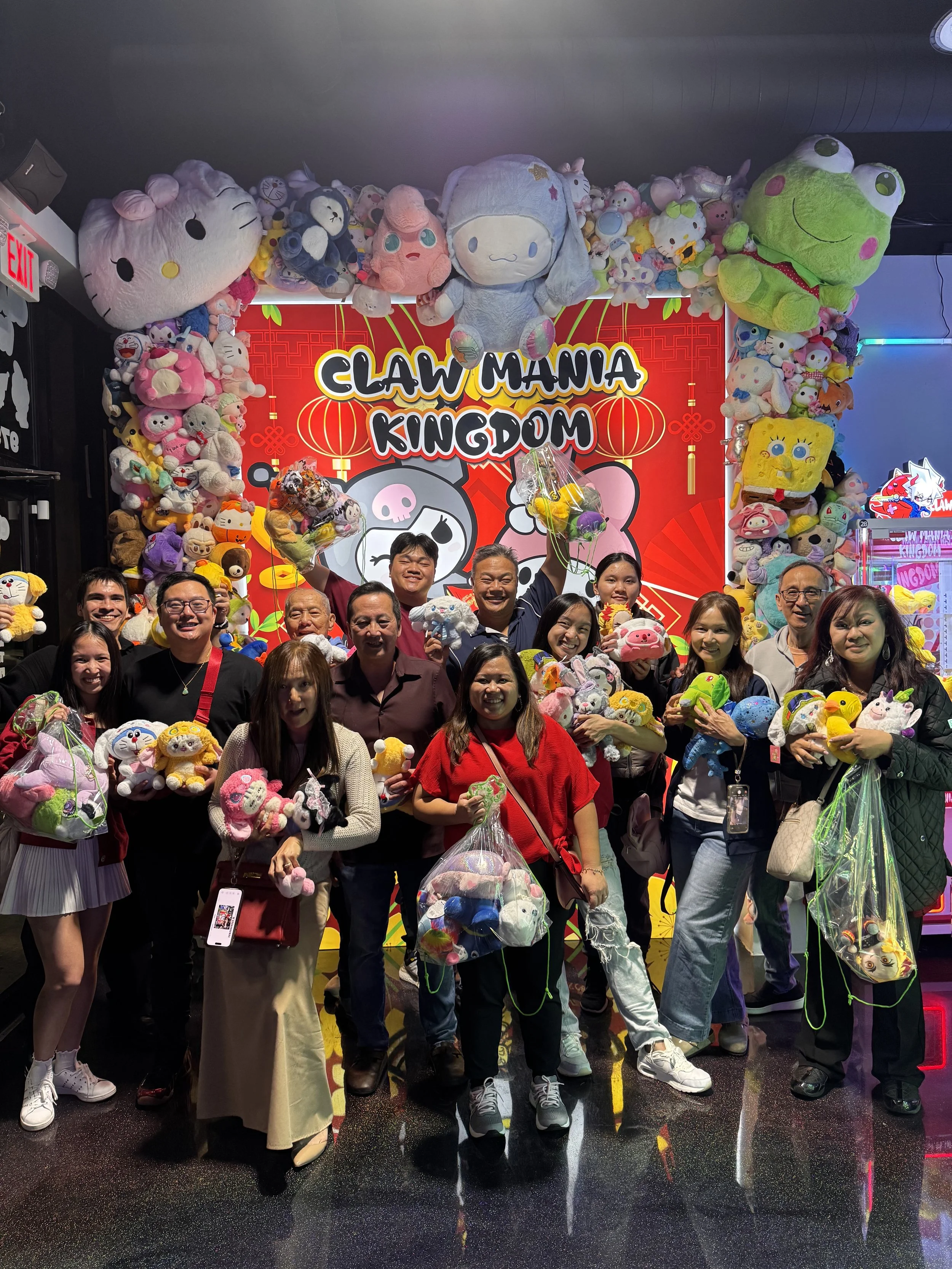 Group of people holding plush toys in front of a large red sign that reads 'Claw Mania Kingdom' decorated with cartoon characters and lanterns, with plush toys arranged around the sign.