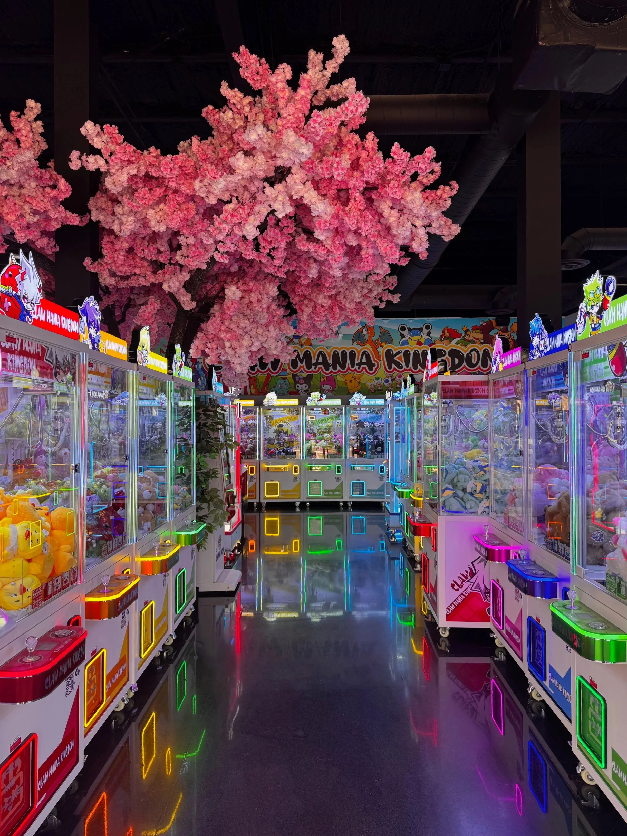 Indoor arcade featuring pink cherry blossom tree canopy overhead, with multiple claw machine arcade games lined up on both sides, illuminated with colorful neon lights.