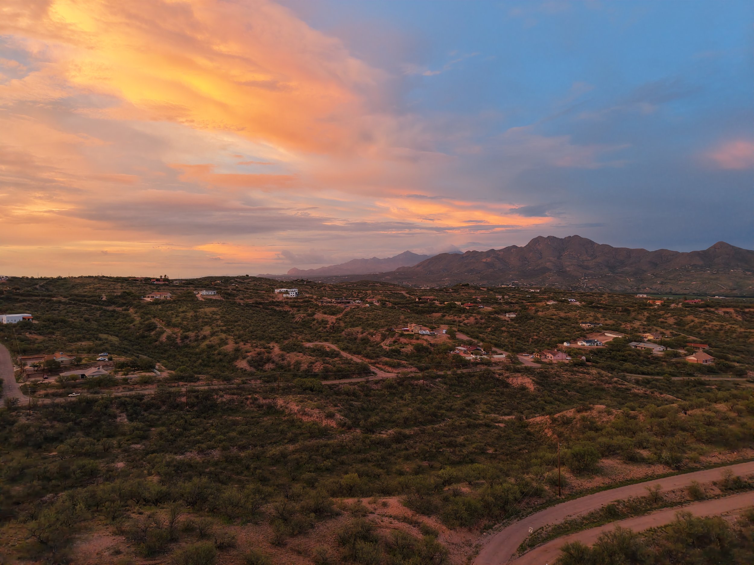 Scenic view of a mountain range under a colorful sunset sky with scattered clouds. A small residential area with houses and a winding road is visible in the foreground.