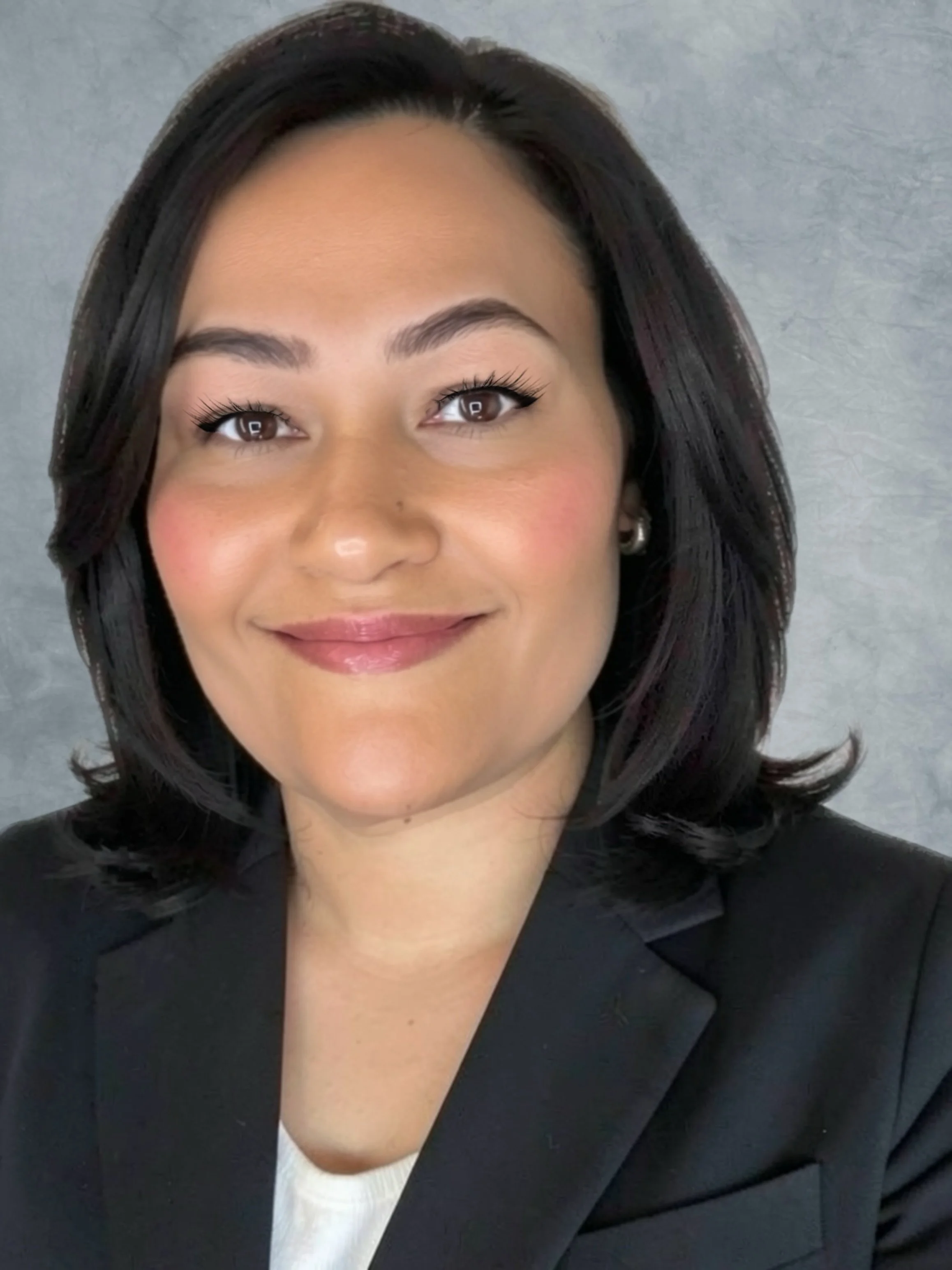 Close-up of a woman with dark hair smiling, wearing a black blazer, white top, and pearl earrings, posing against a plain gray background.