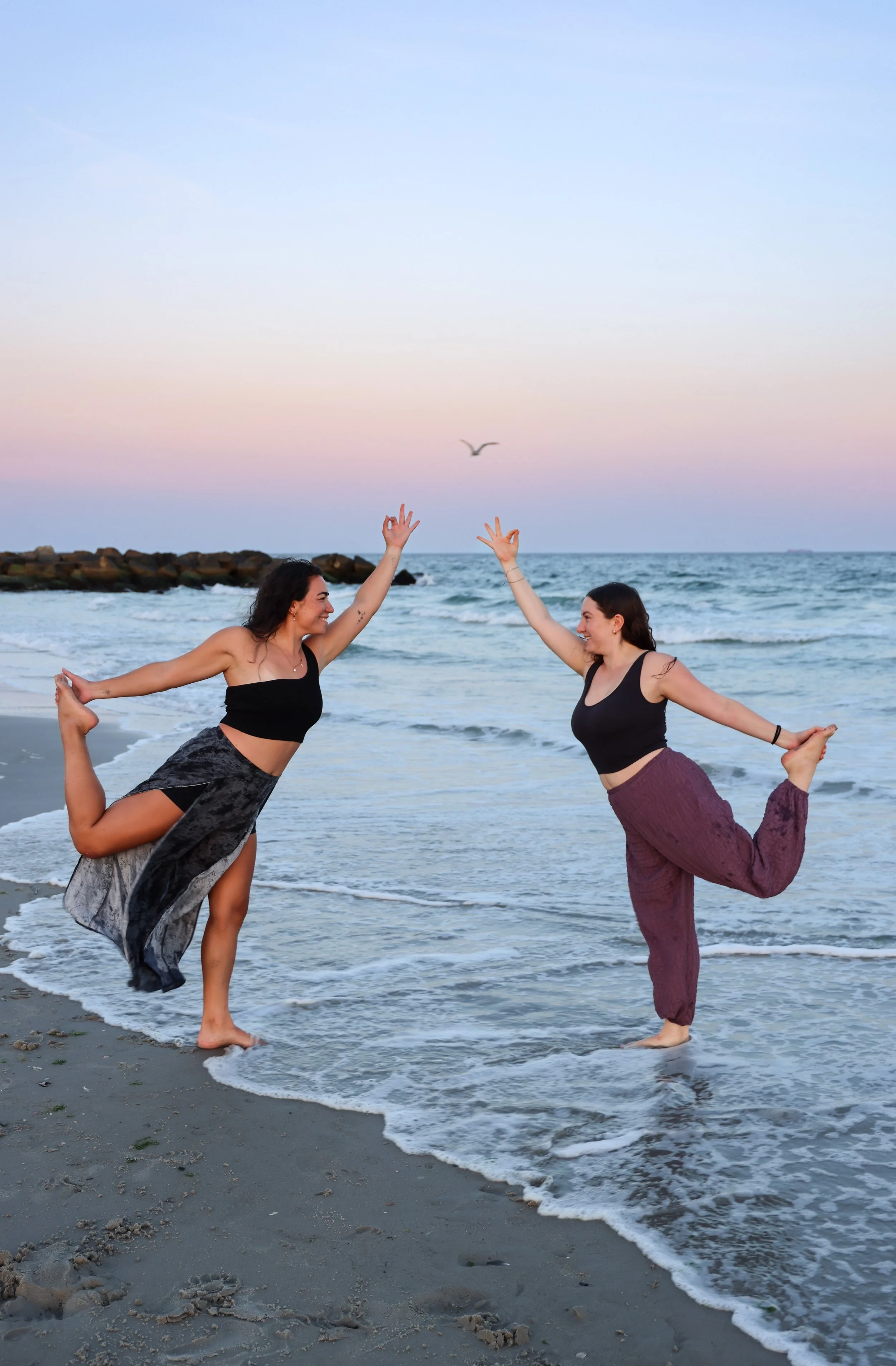 Two women practicing yoga on the beach at sunset, balancing on one leg with arms raised, near the ocean waves.