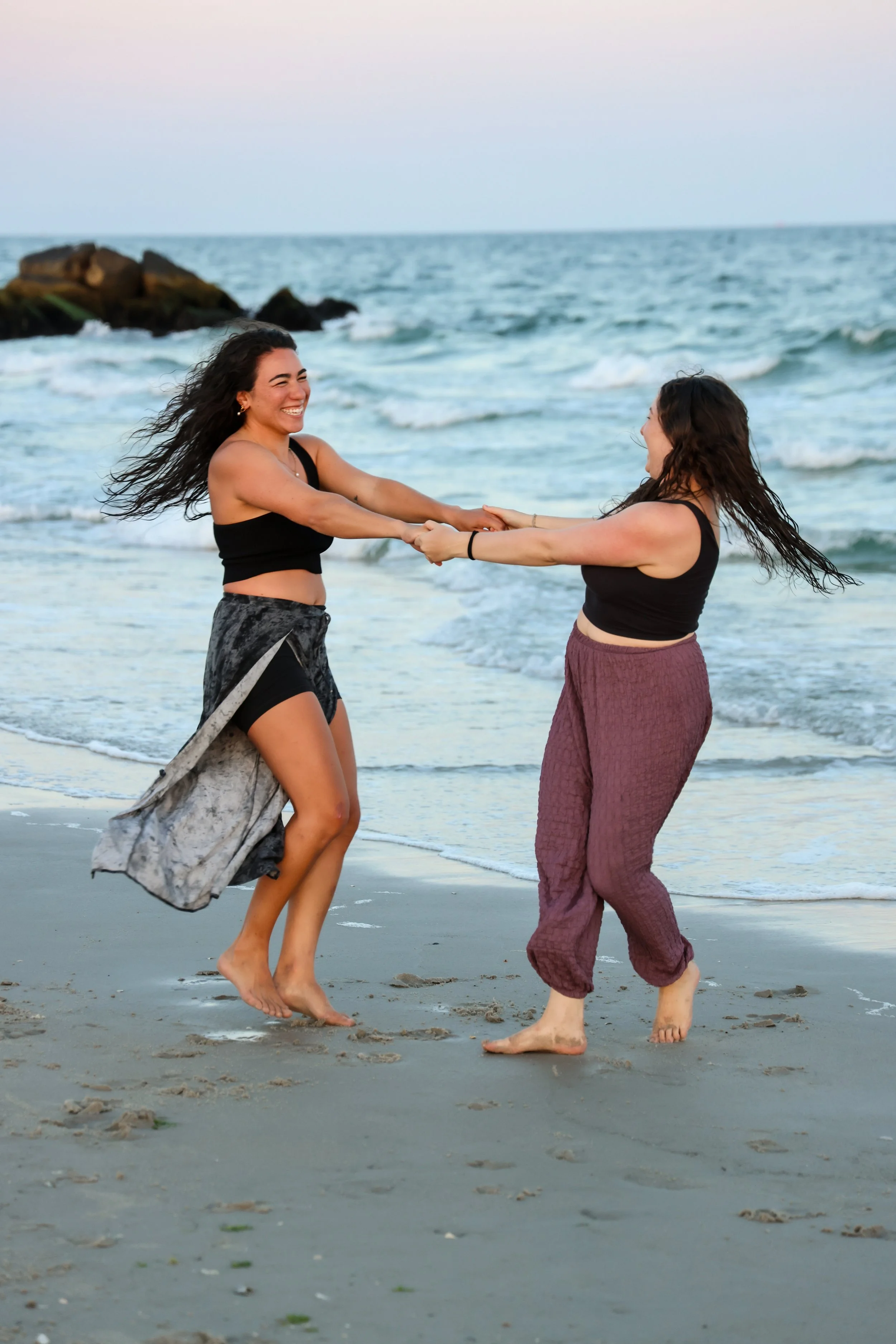 Two women with long hair and welcoming smiles hold hands and dance on the beach near the ocean with waves and rocks in the background. They are barefoot and dressed in casual summer clothing.