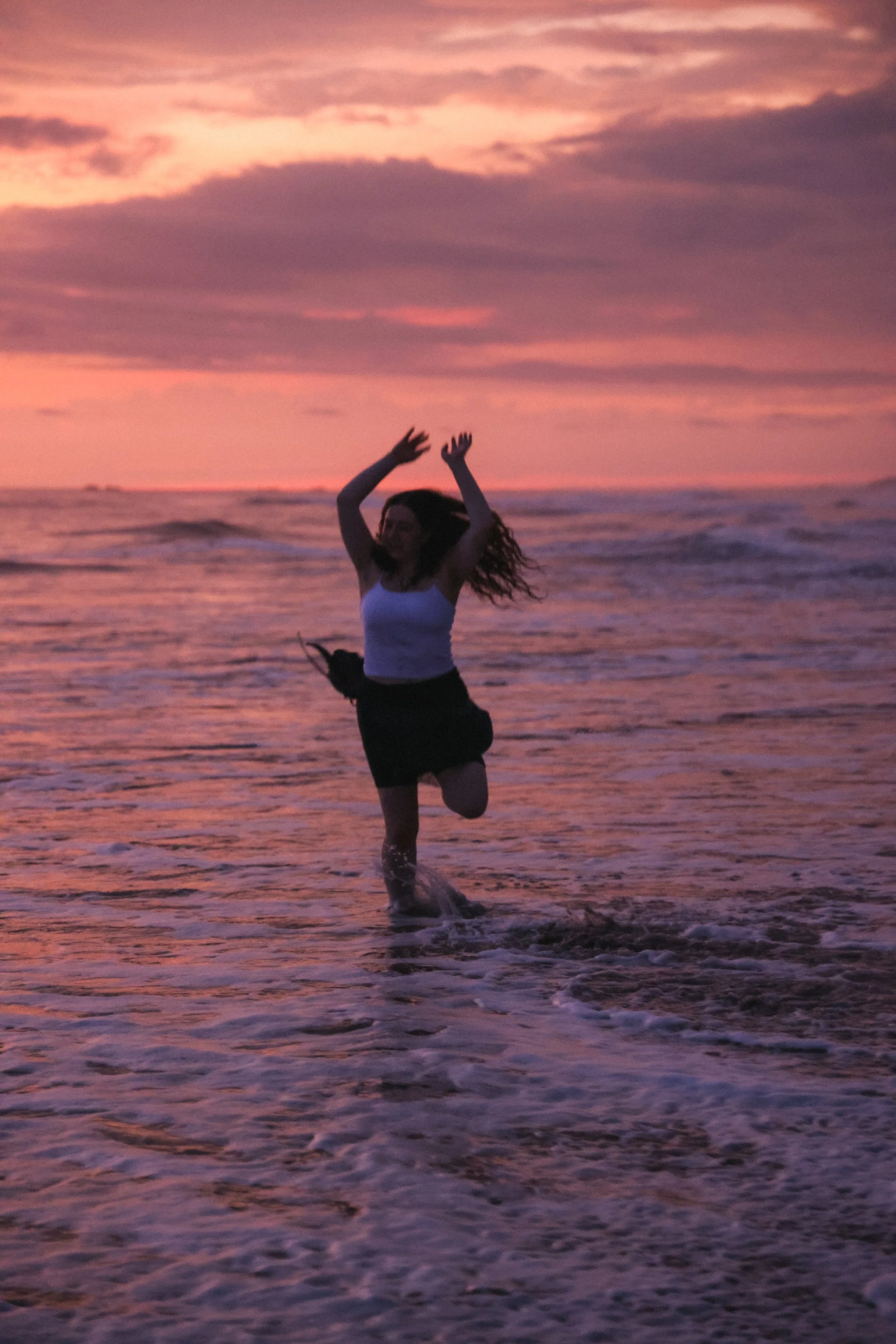 A woman with curly hair wearing a white tank top and black shorts jumping in the ocean at sunset, with purple and pink sky and waves around her.