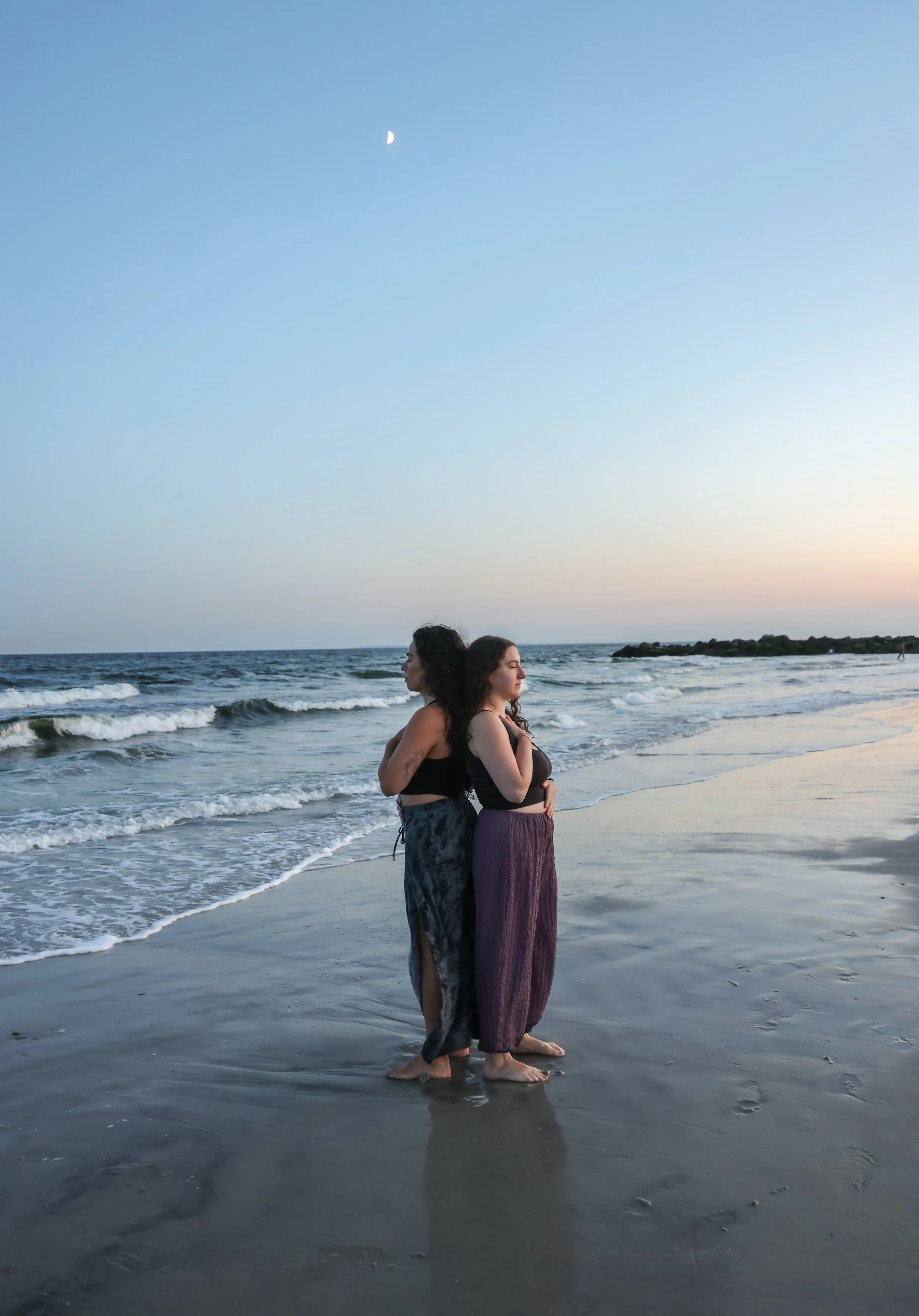 Two women standing side by side on the beach at sunset, facing opposite directions with eyes closed, wearing black tops and long skirts, with the ocean waves in the background and a clear sky with a visible moon.