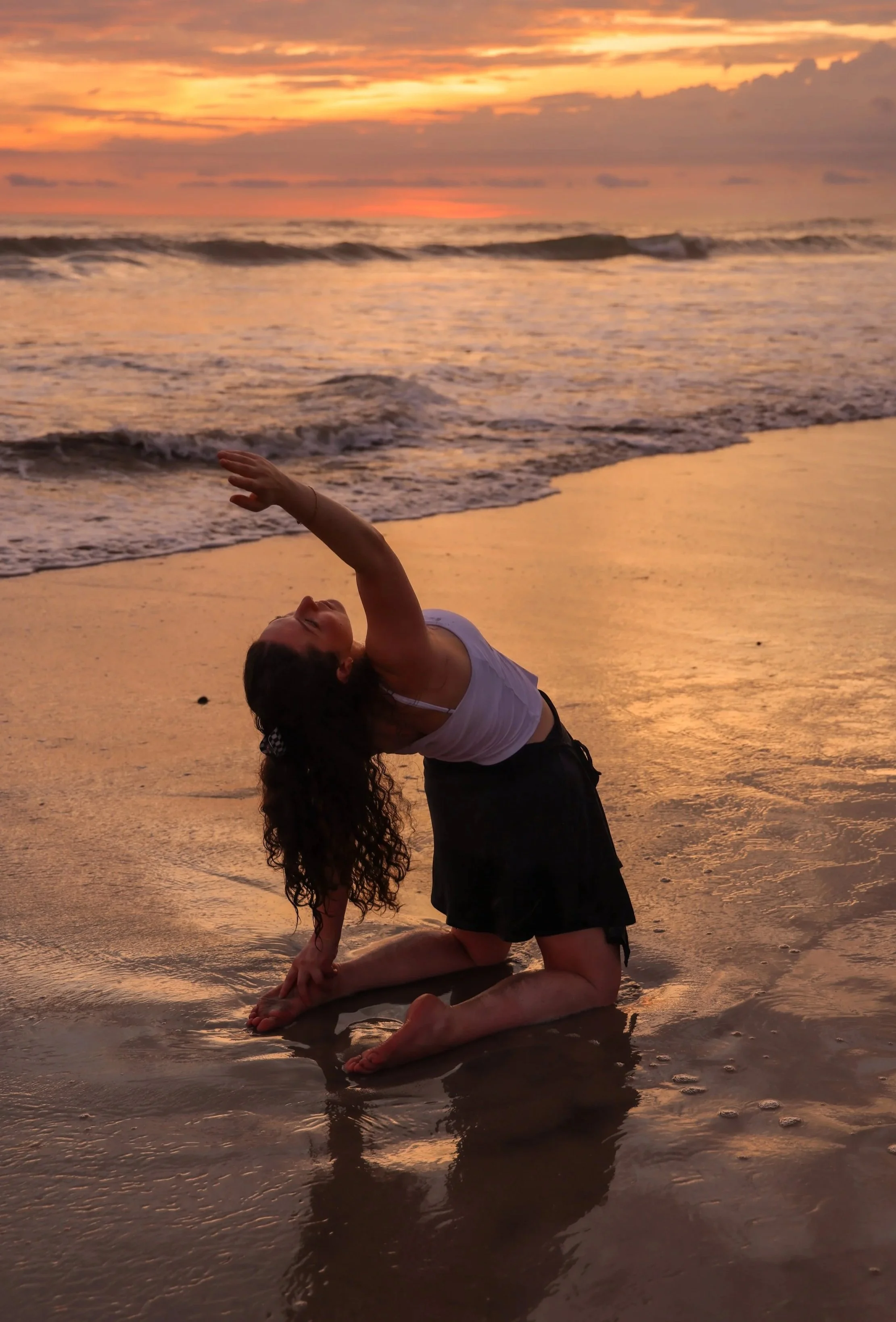A woman practicing yoga on a beach at sunset, kneeling on the sand with her body arched backward and one arm reaching upward, with the ocean waves and a colorful sky in the background.