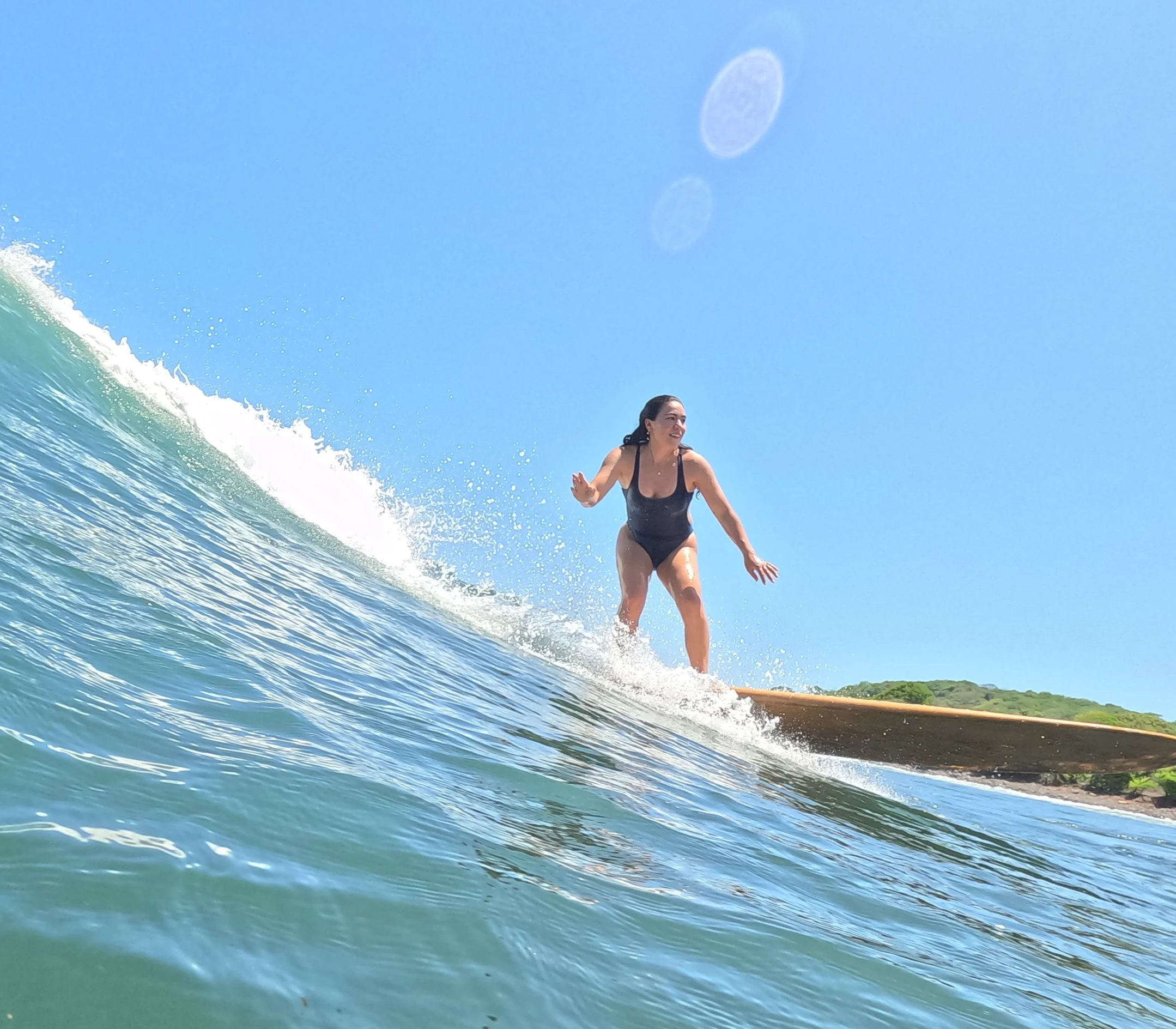A woman in a black swimsuit surfing on a wave with a bright blue sky and a distant hillside.