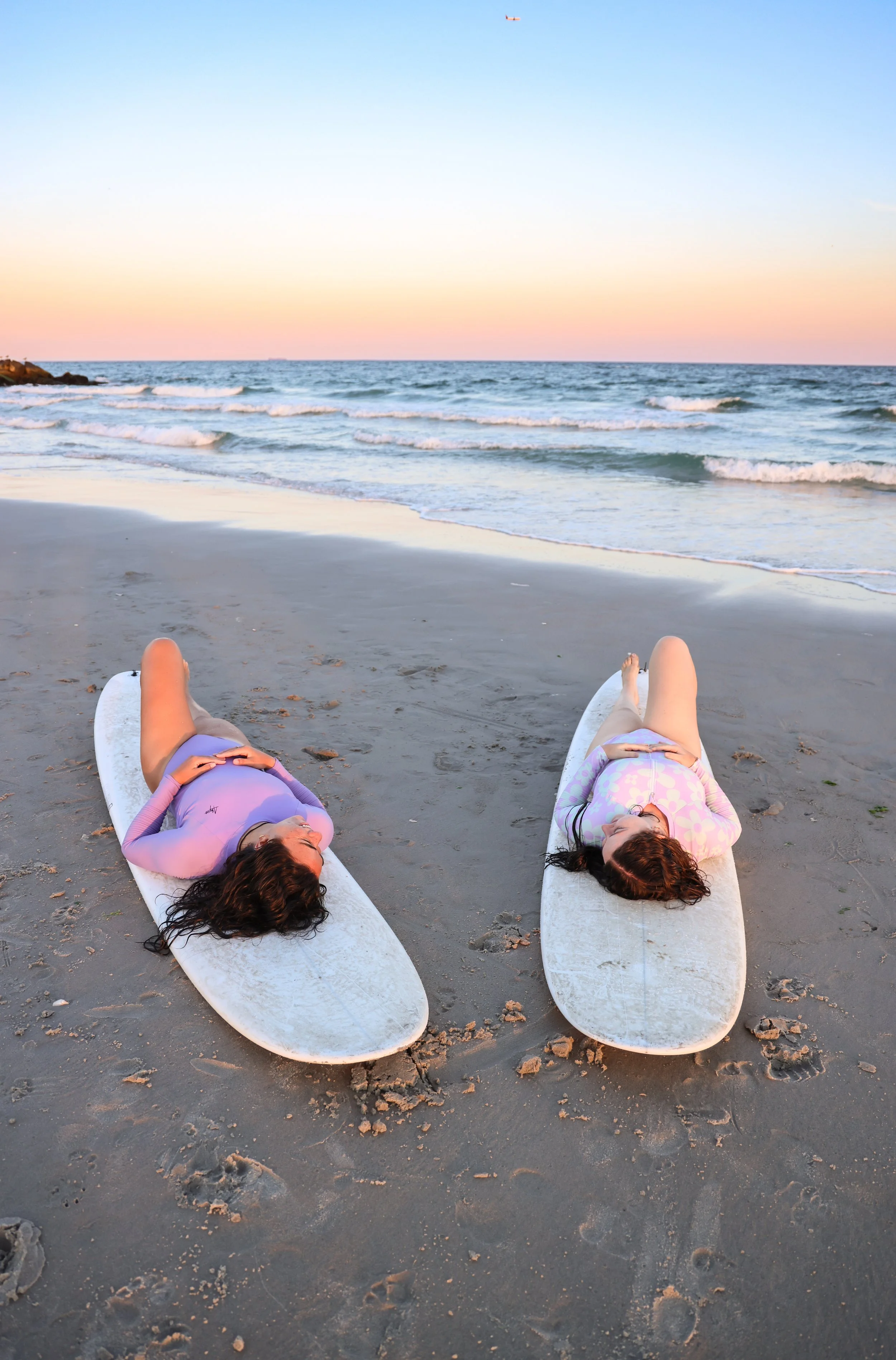 Two women lying on surfboards on a sandy beach, facing the ocean at sunset, with waves and a pastel-colored sky in the background.