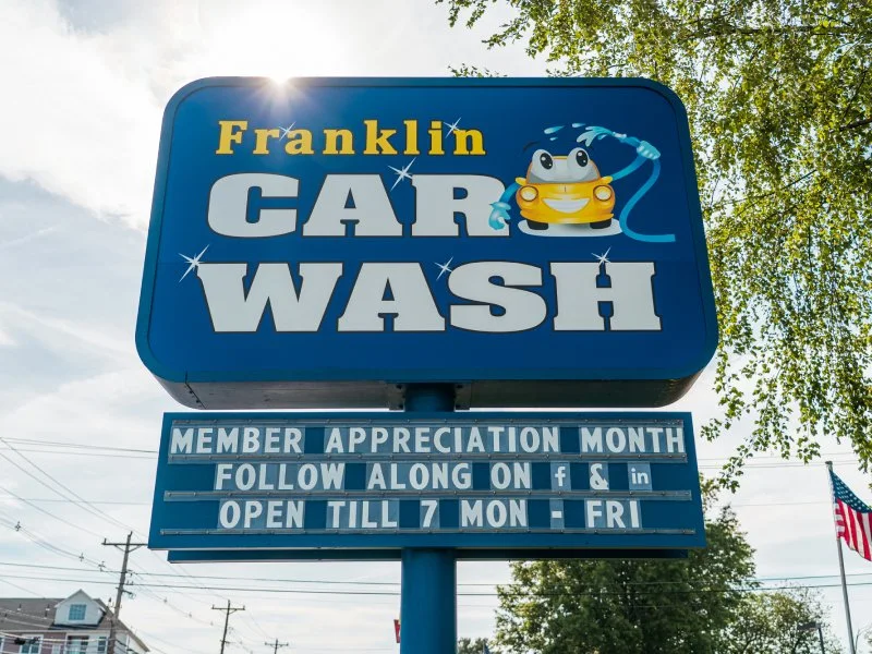 Blue sign for Franklin Car Wash with cartoon frog washing a yellow car, and a message about Member Appreciation Month and hours of operation.